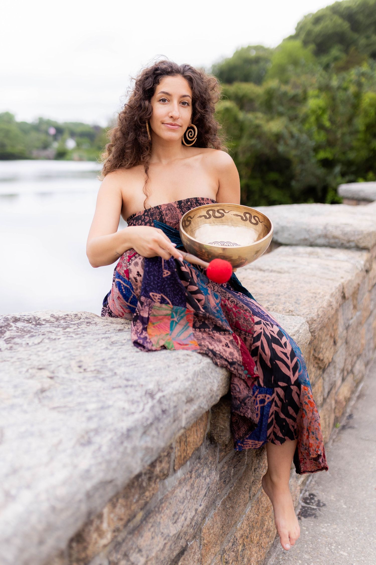 Person in floral dress sits on stone wall holding meditation bowl near waterfront.