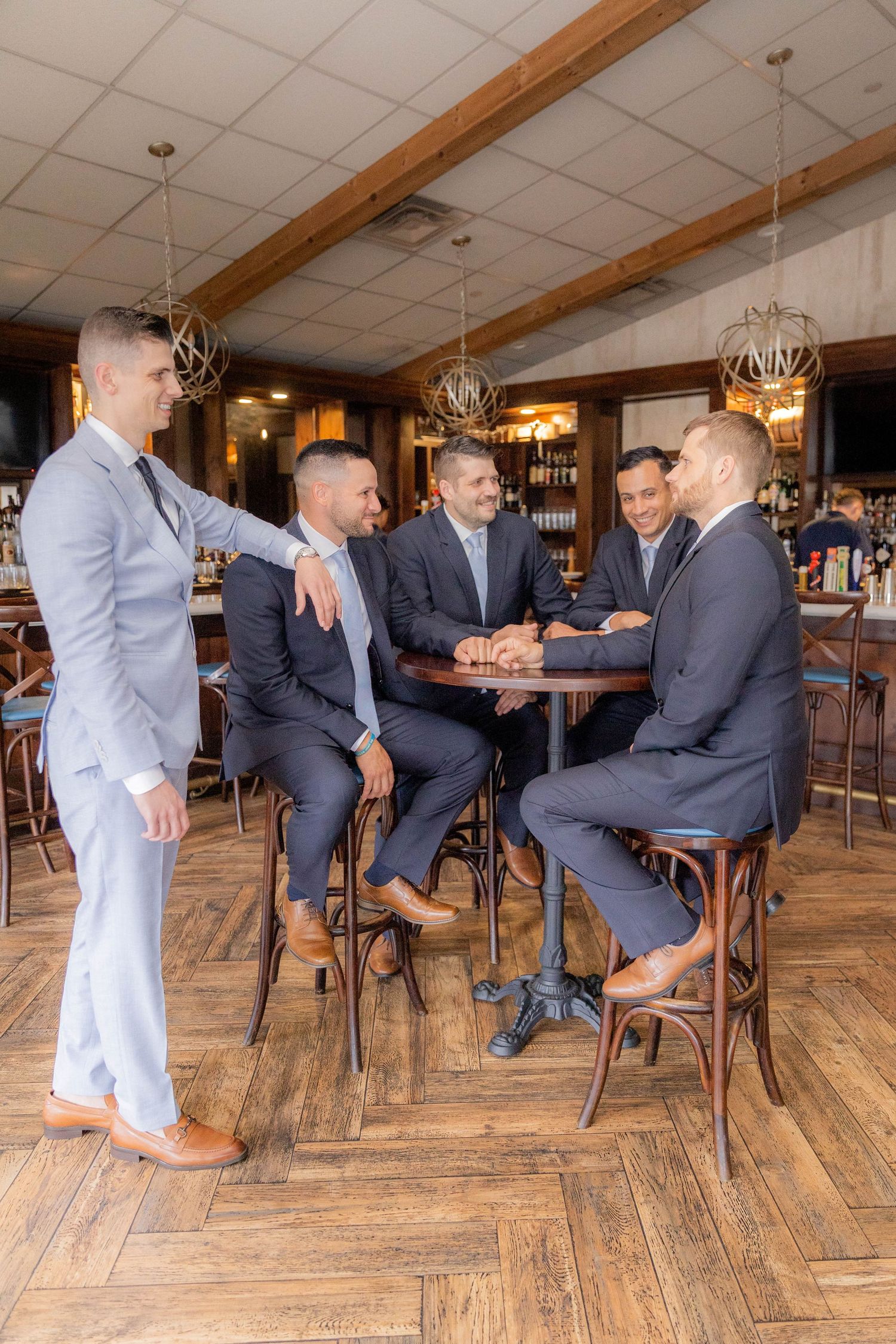 A group of men in suits gather around a high-top table at an upscale bar with rustic wood floors and pendant lighting.