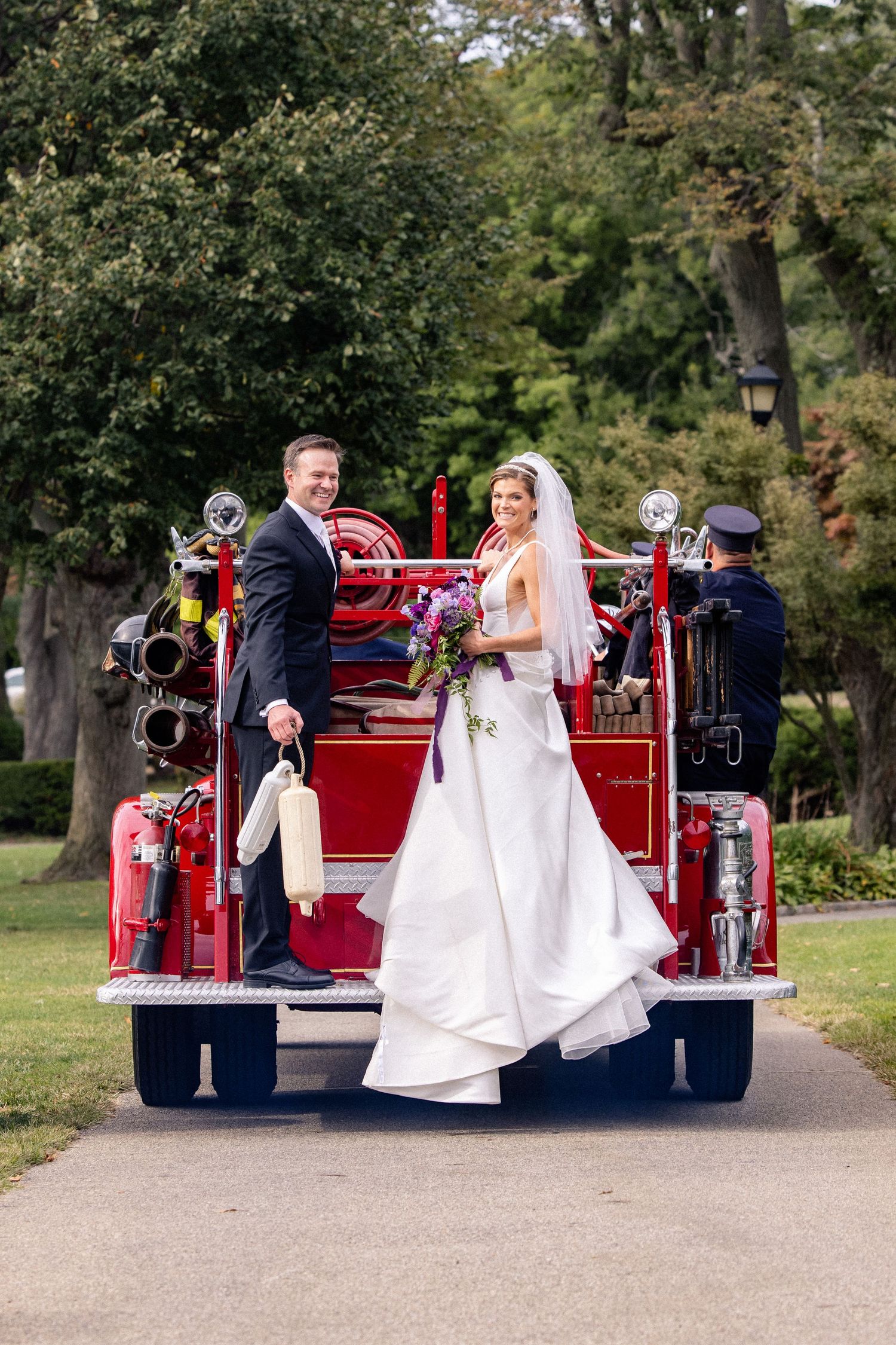 A couple in wedding attire poses on the back of a classic red fire engine in a park setting.