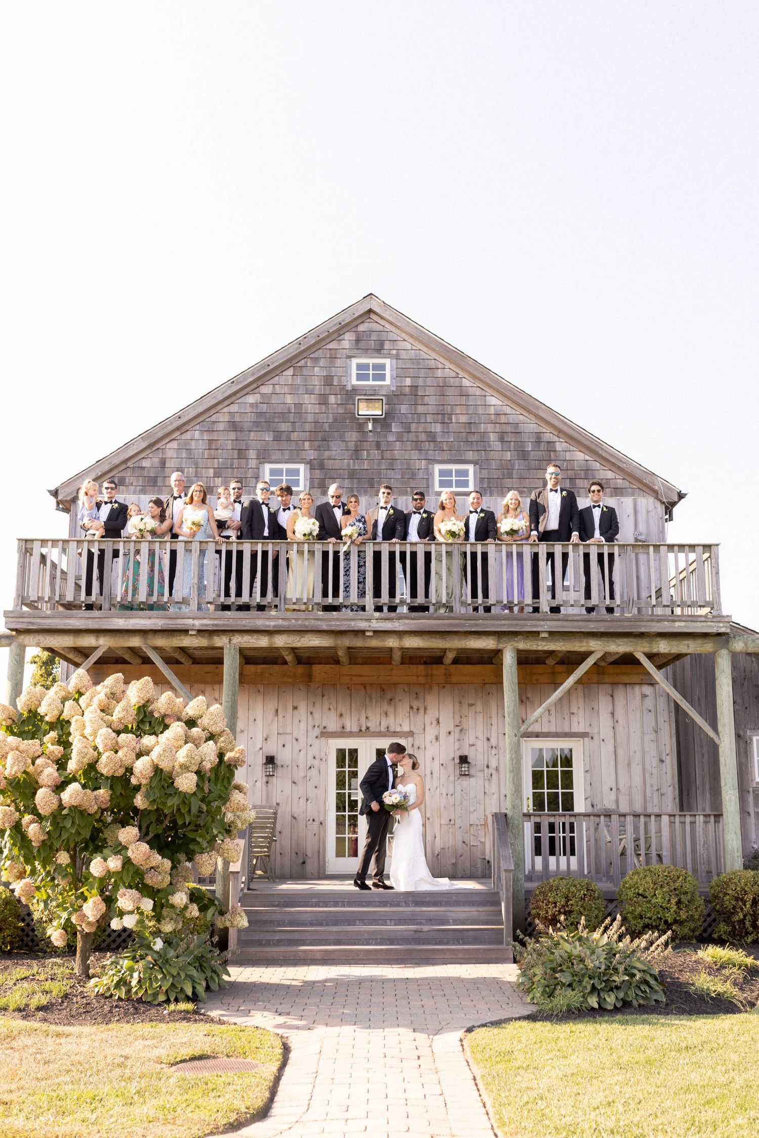 Wedding party stands on rustic barn balcony decorated with cream colored flowers at sunset.