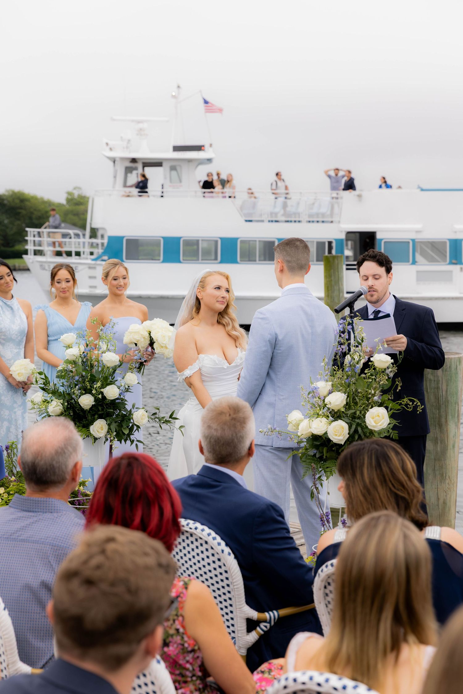 Wedding guests gather on a dock for an outdoor ceremony with white floral arrangements and boats in view.