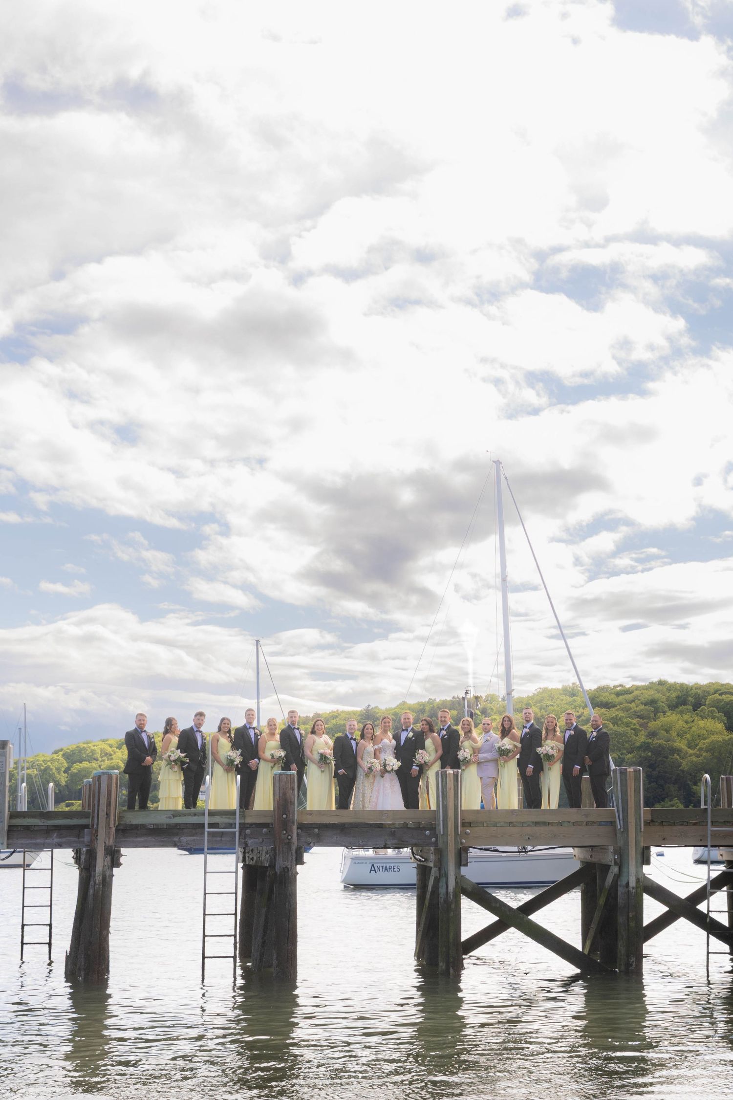 A wedding party stands in formal attire along a wooden dock with boats and water in the background on a cloudy day.