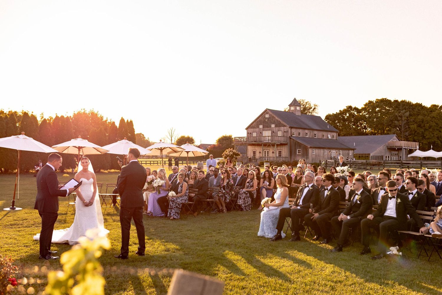 Wedding guests gather outdoors at a rustic farm venue during golden hour with tents and vintage decor in view.