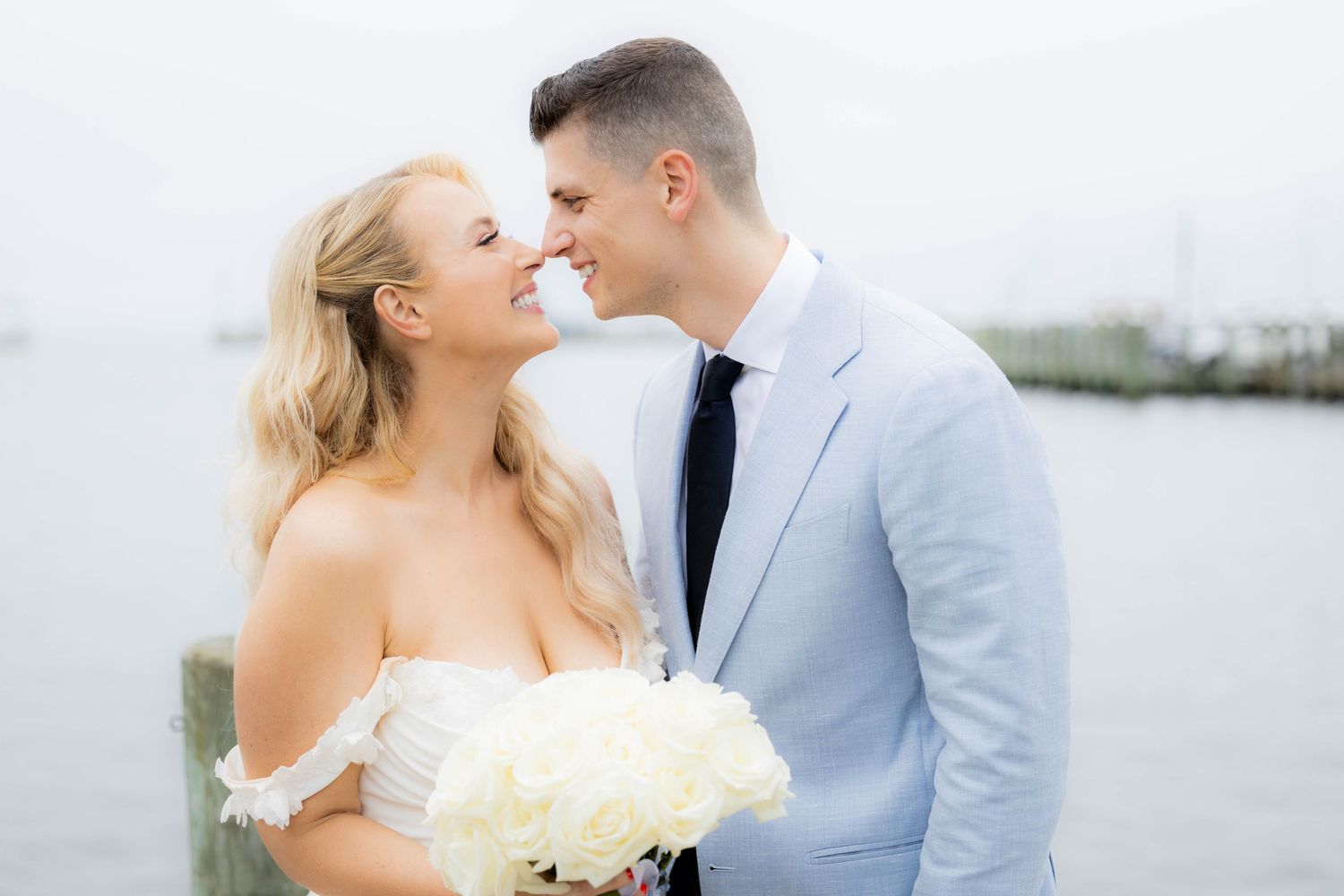A couple in wedding attire shares romantic moments by the waterfront in a light gray and white color scheme.
