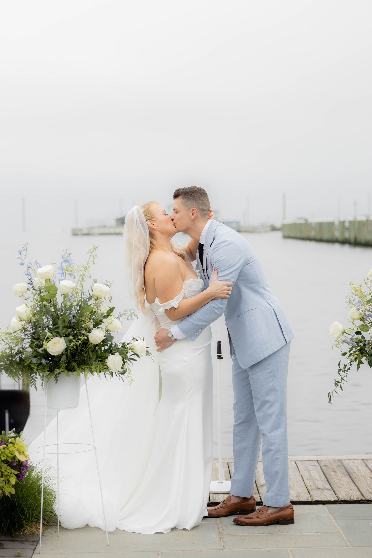 A romantic wedding couple shares intimate moments on a waterfront dock decorated with elegant white floral arrangements.