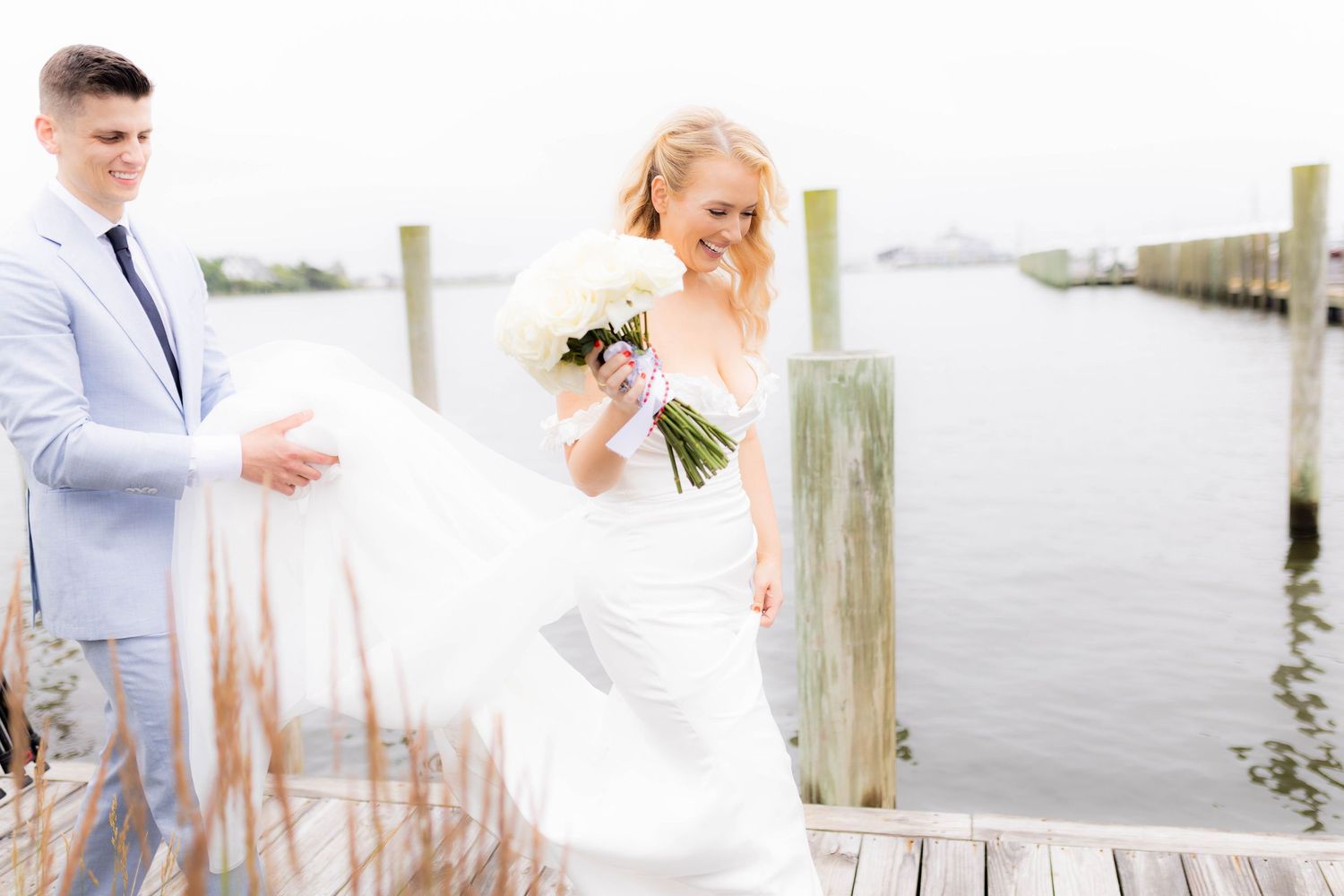 A bride and groom share romantic moments along a waterfront dock in soft gray and white tones.