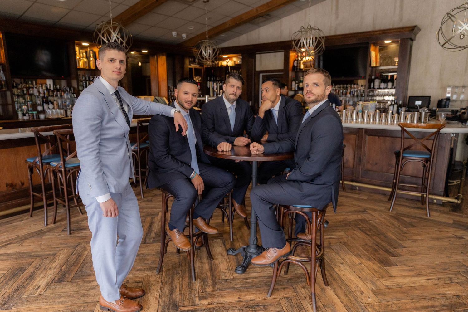 Groomsmen in gray suits share drinks and conversation at a rustic wooden bar.