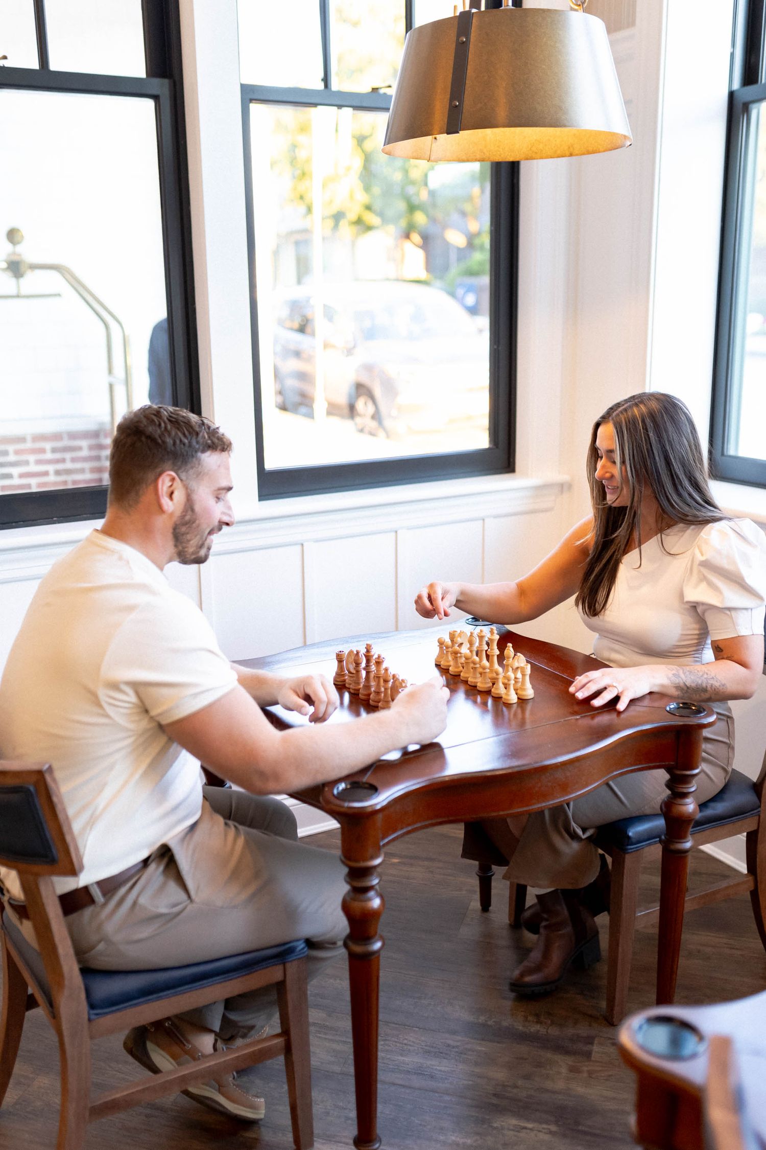 A couple plays chess together at a wooden table in a bright indoor space with large windows.