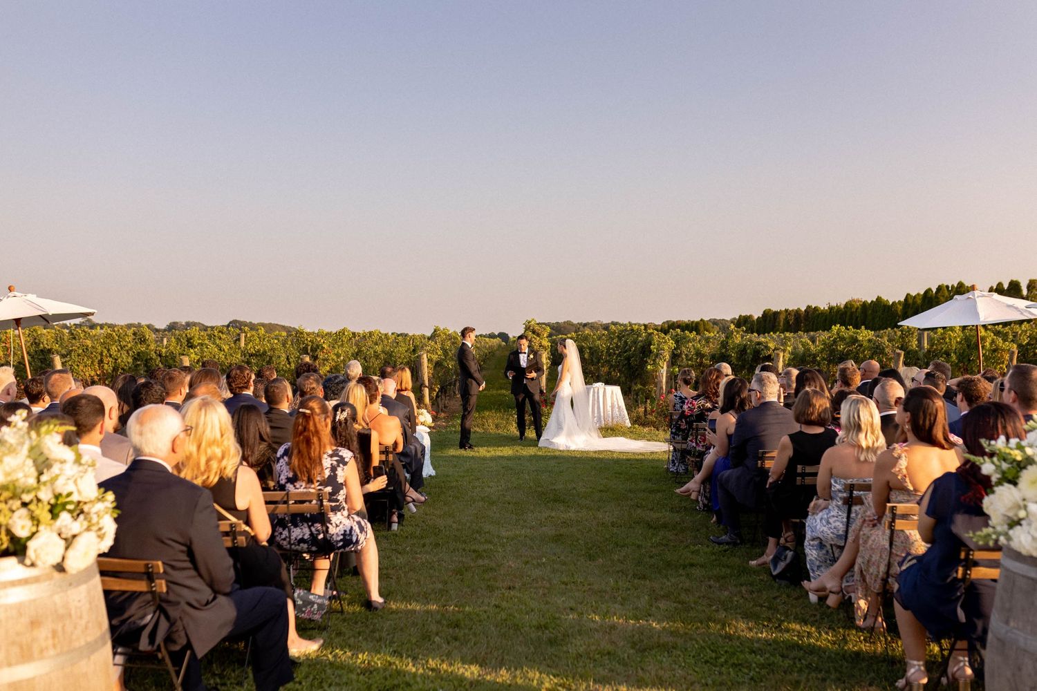 A wedding ceremony takes place on a grassy hill during golden hour with guests seated in white chairs.