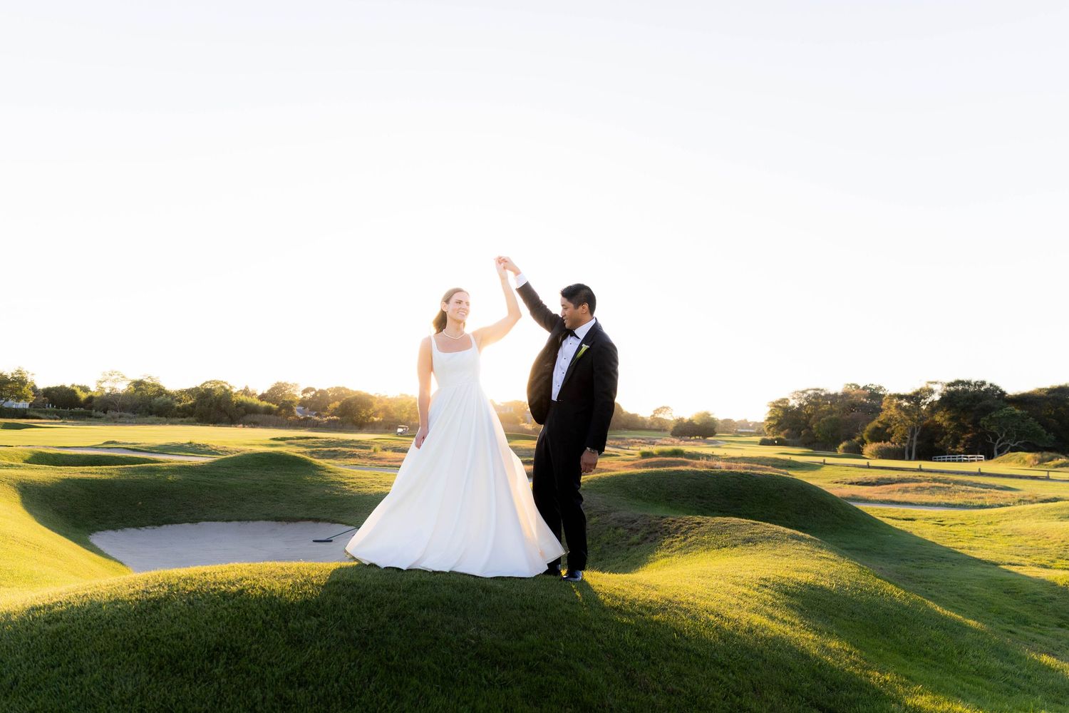 Newlyweds share a joyful moment on a golf course with sand traps at sunset.