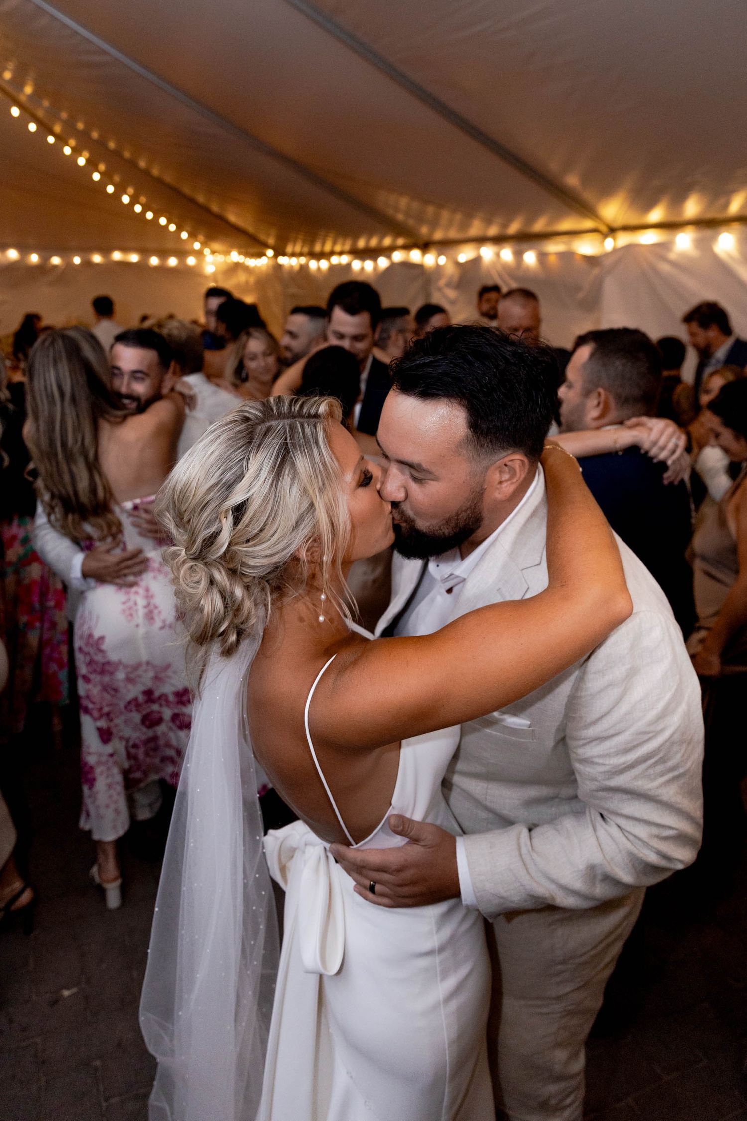 A romantic moment on the dance floor at a wedding reception under a tent with string lights overhead.