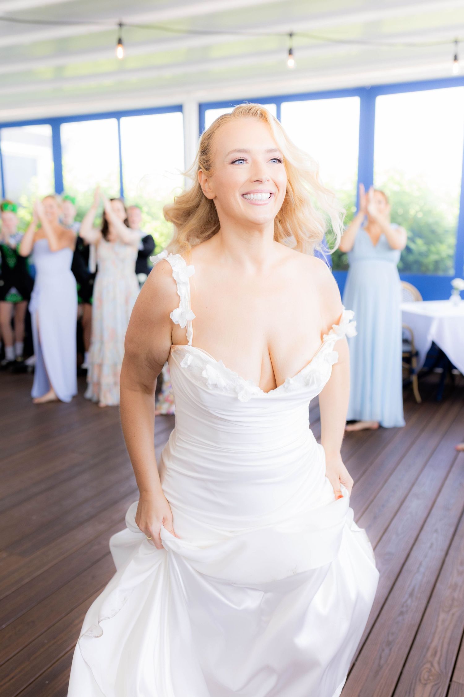 A bride in a white wedding dress dances joyfully on a wooden deck with guests in the background.