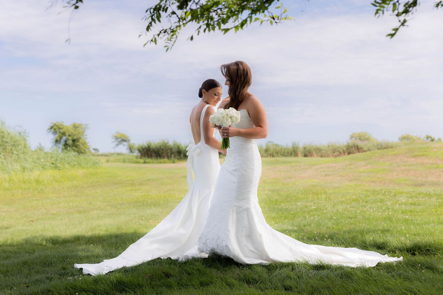 Two brides in elegant white wedding gowns embrace on a grassy hilltop with blue sky and greenery in the background.
