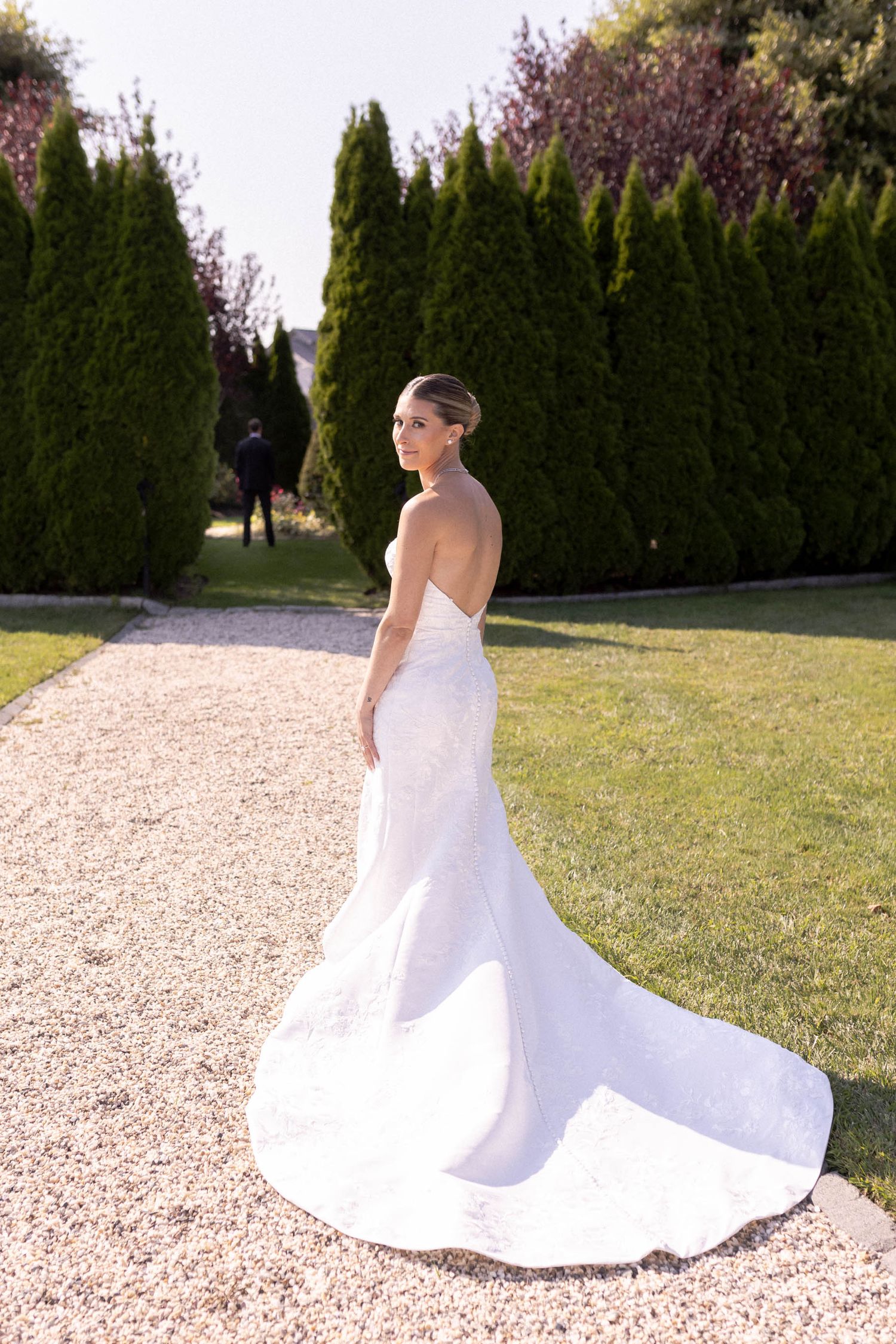 A flowing white wedding dress with a long train is displayed on a gravel path surrounded by green landscaping.