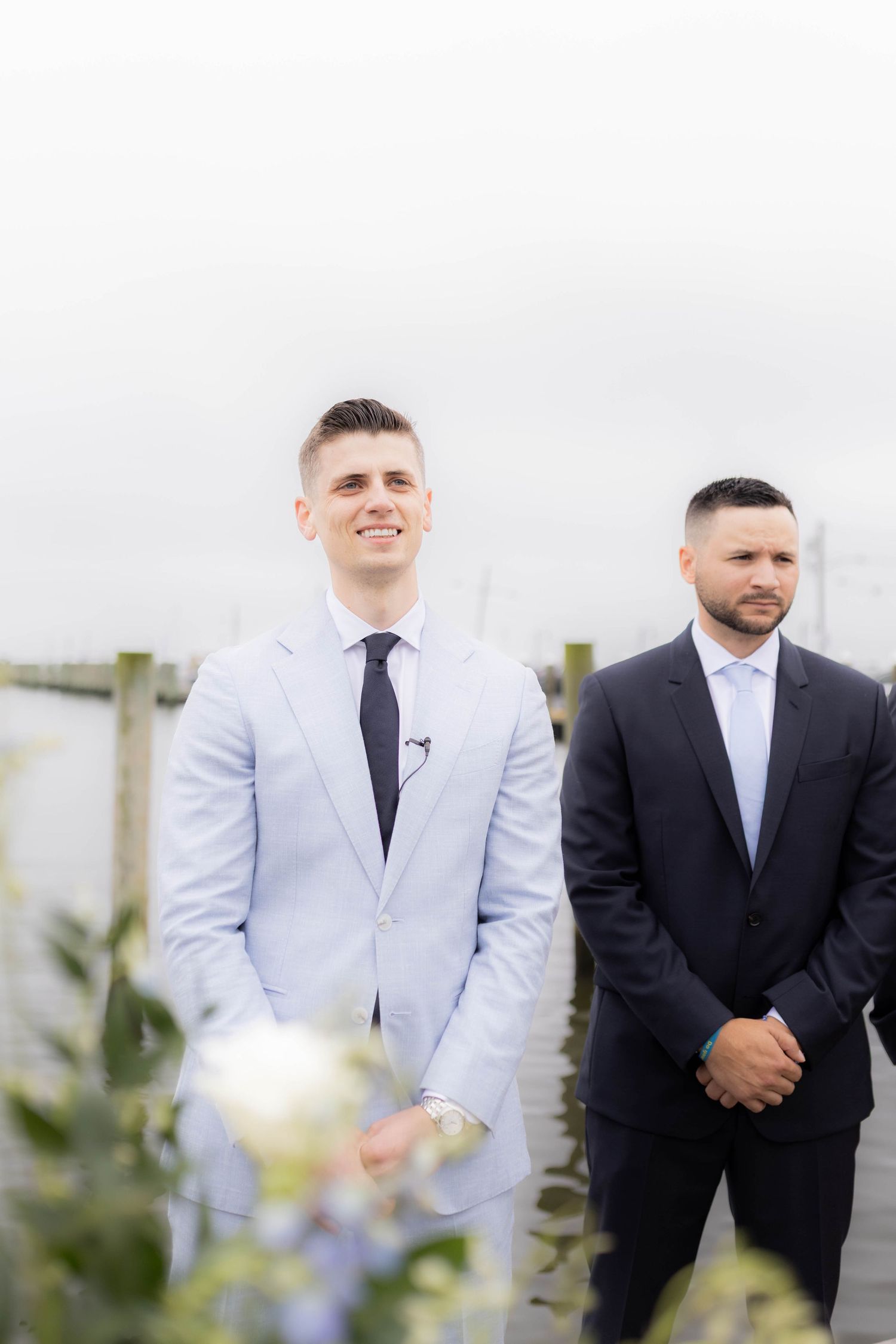 Two men in formal attire stand together smiling at an outdoor wedding ceremony.