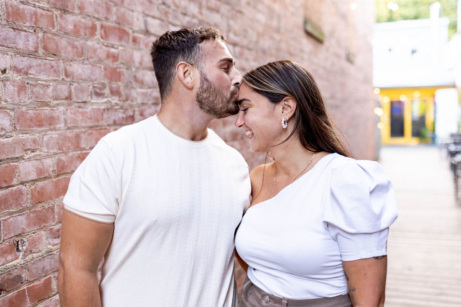 Romantic couple in white shirts sharing an intimate moment against a brick wall outdoors.
