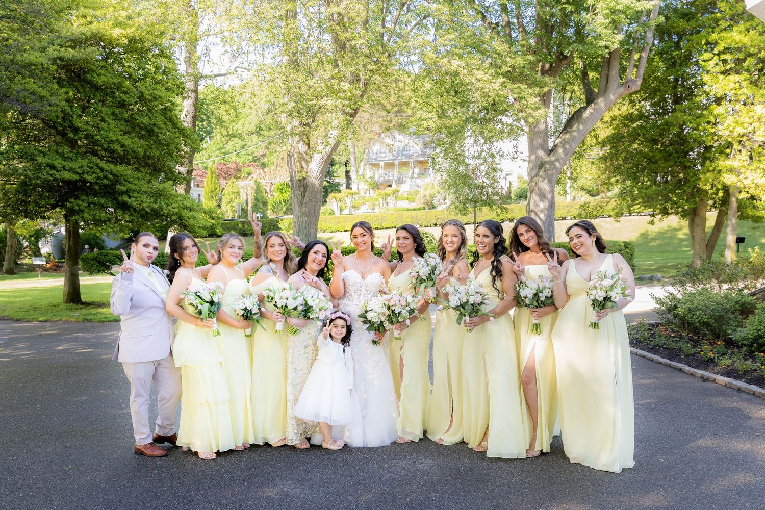 Wedding party members in pastel yellow and white dresses pose together on a tree-lined pathway.