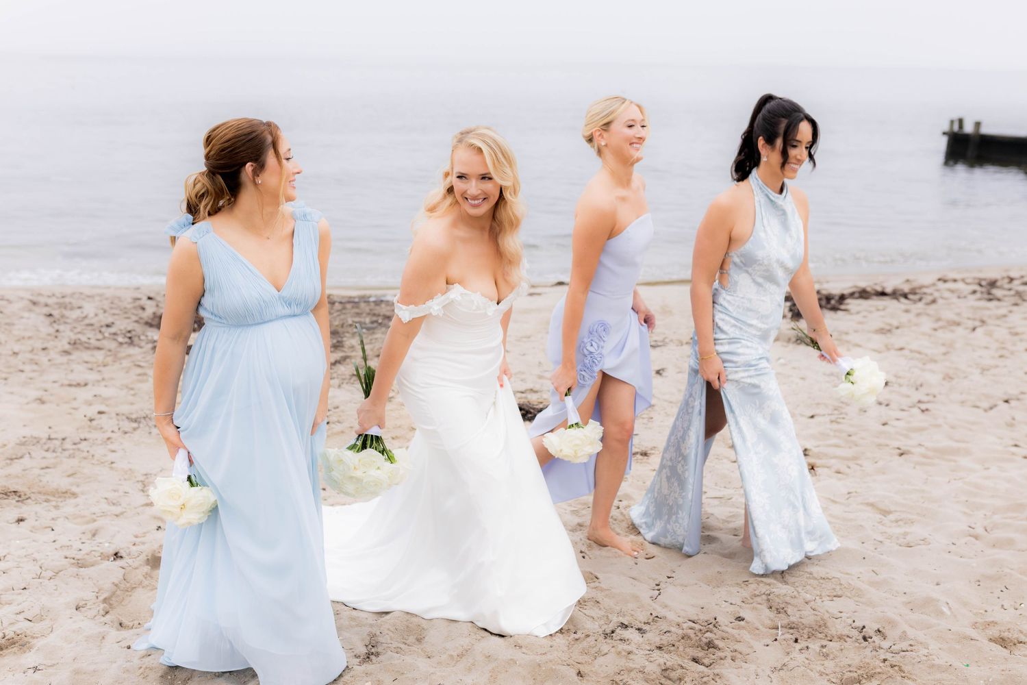 Bridesmaids in light blue dresses walk along a sandy beach with a bride in a white wedding gown.