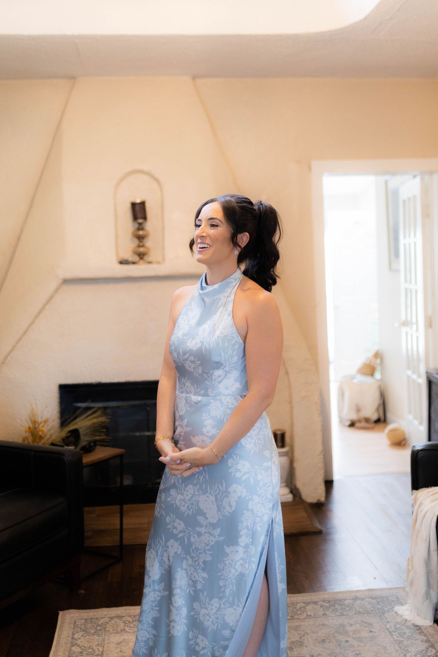 A person in a light blue floral halter dress stands smiling in a bright living room with a fireplace.