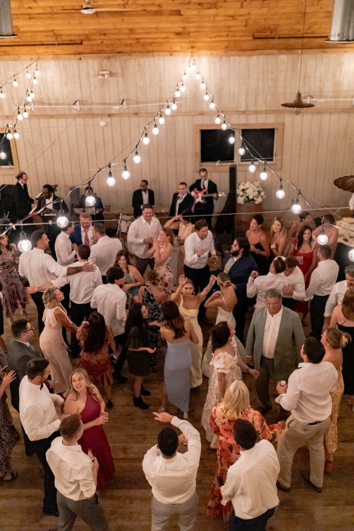 Wedding guests dancing on a rustic wooden dance floor under string lights at an indoor reception venue.