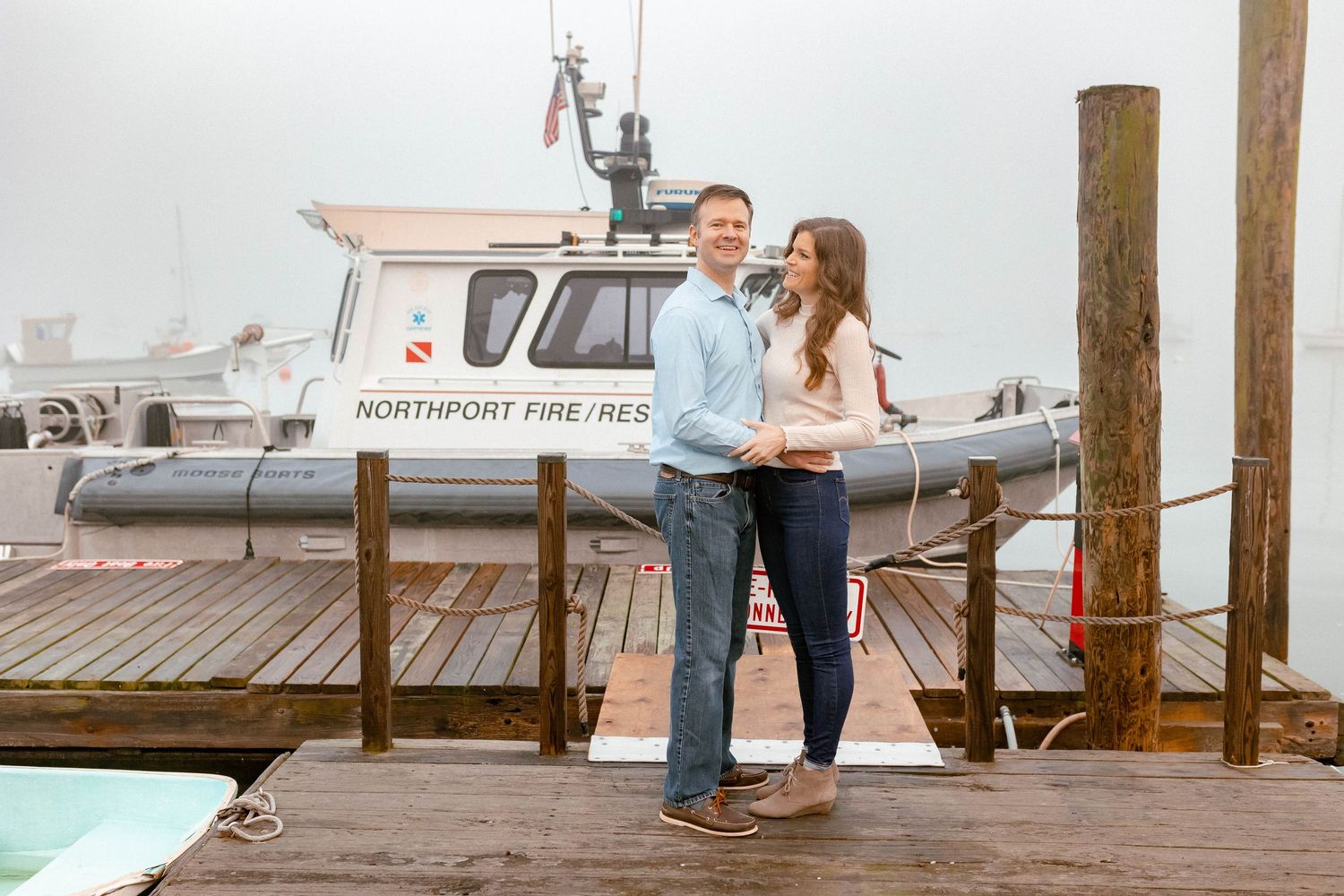 Couple stands together on a wooden dock with boats and water in the background.