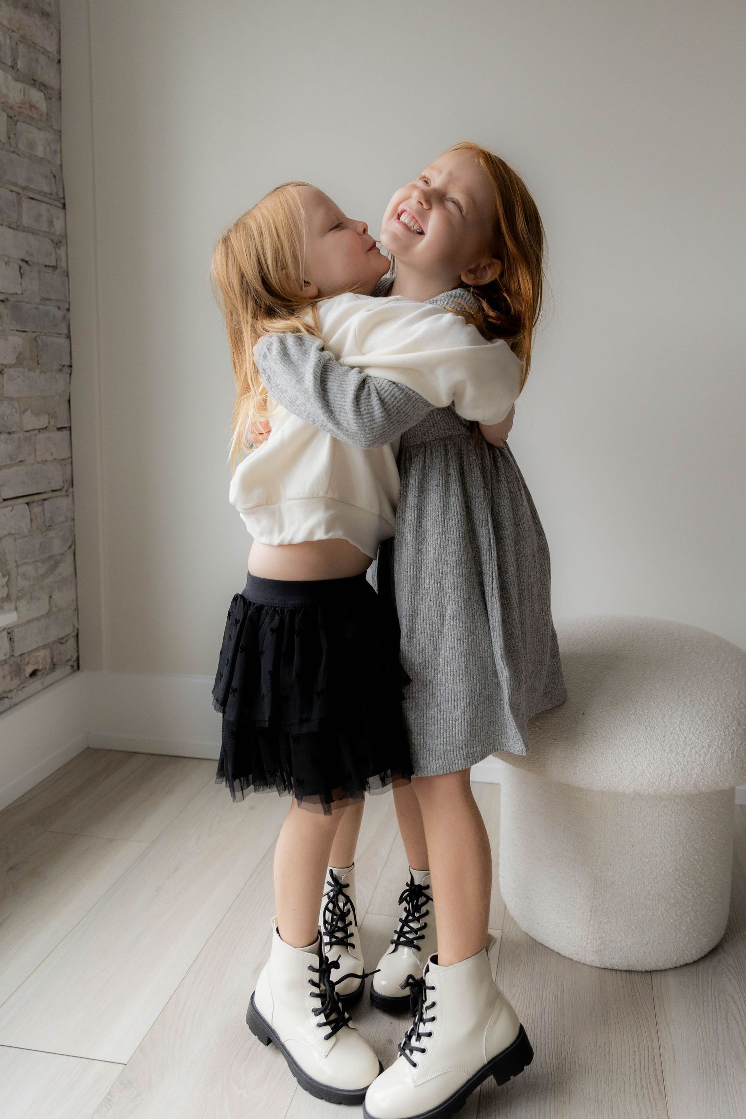 Two young children in matching white boots share a playful moment on a light wood floor. Huntington Loft. Fireside Sparrow Photography
