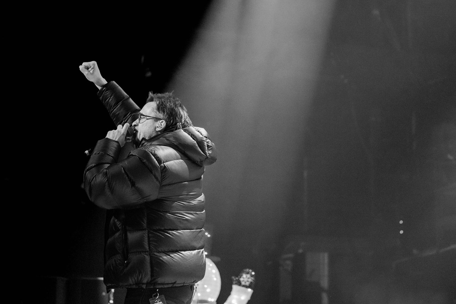 Dramatic black and white concert photo with performer illuminated by spotlight on stage.