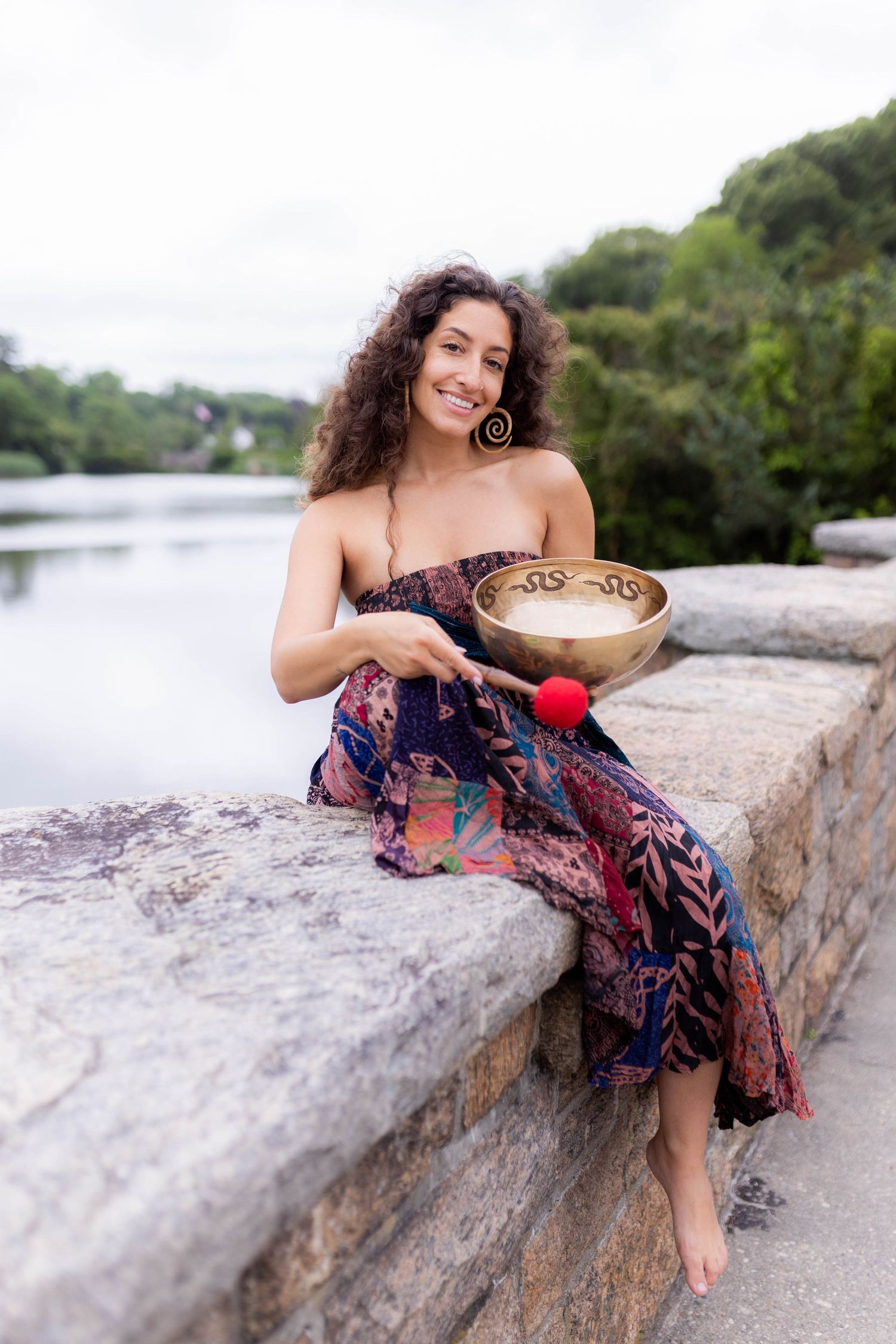 Person in patterned dress holds singing bowl while sitting on stone wall near water.
