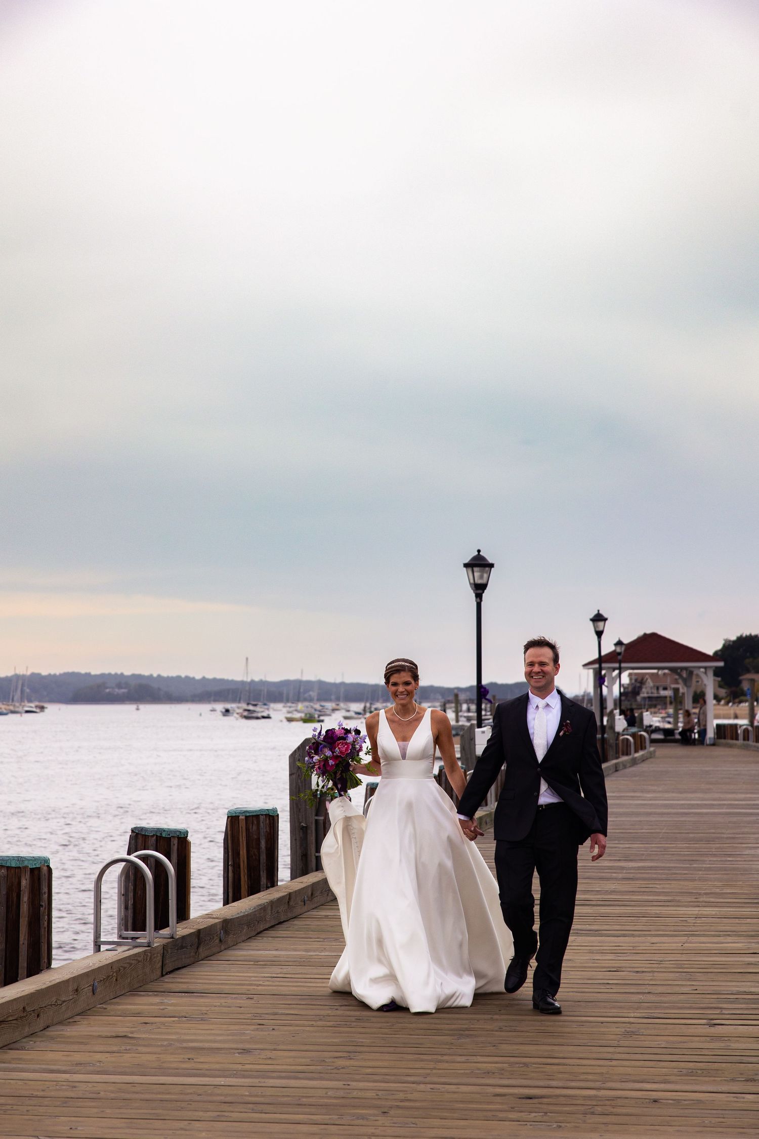 A couple holding hands and smiling while walking on a wooden pier with boats and water in the background.