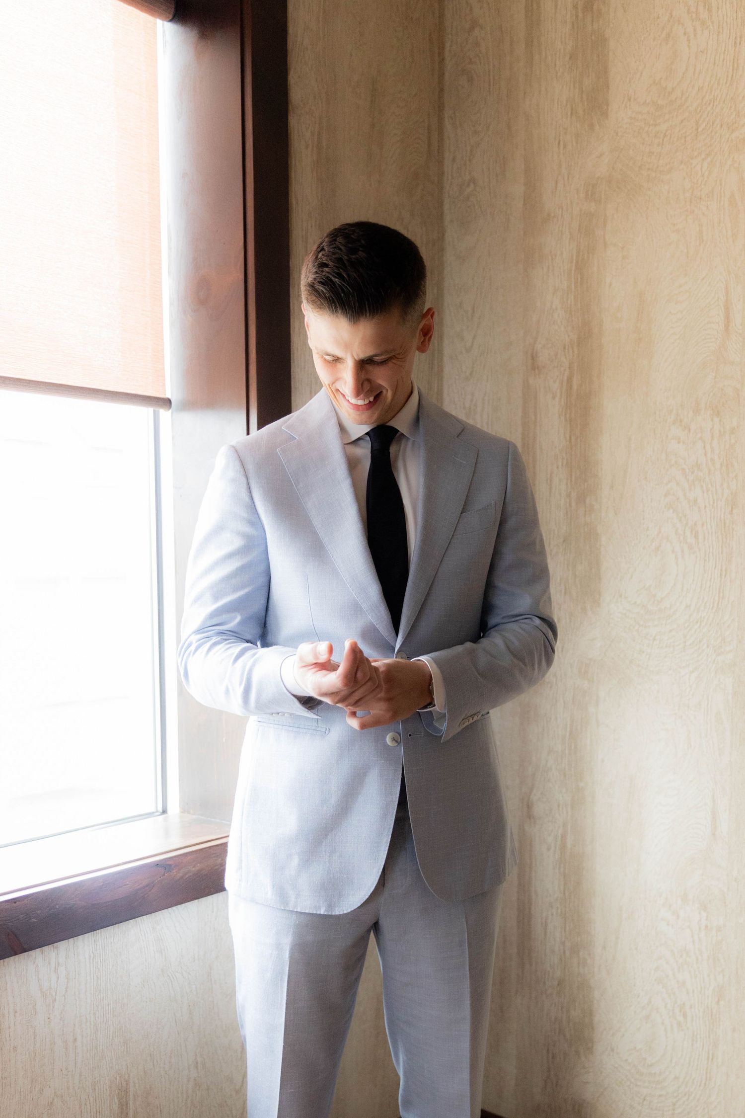A person in a light gray suit stands near a window looking down at their phone.