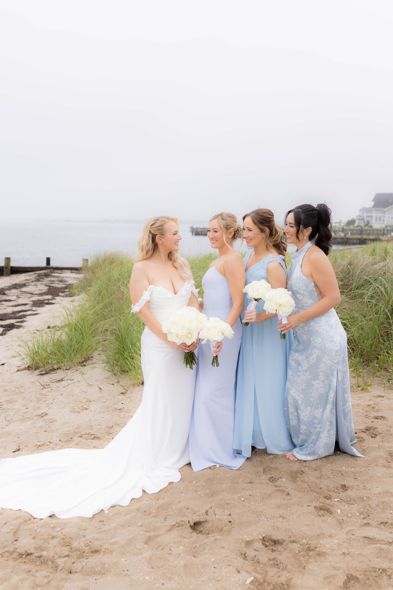 Bridesmaids in light blue dresses gather with the bride on a sandy beach for wedding photos.