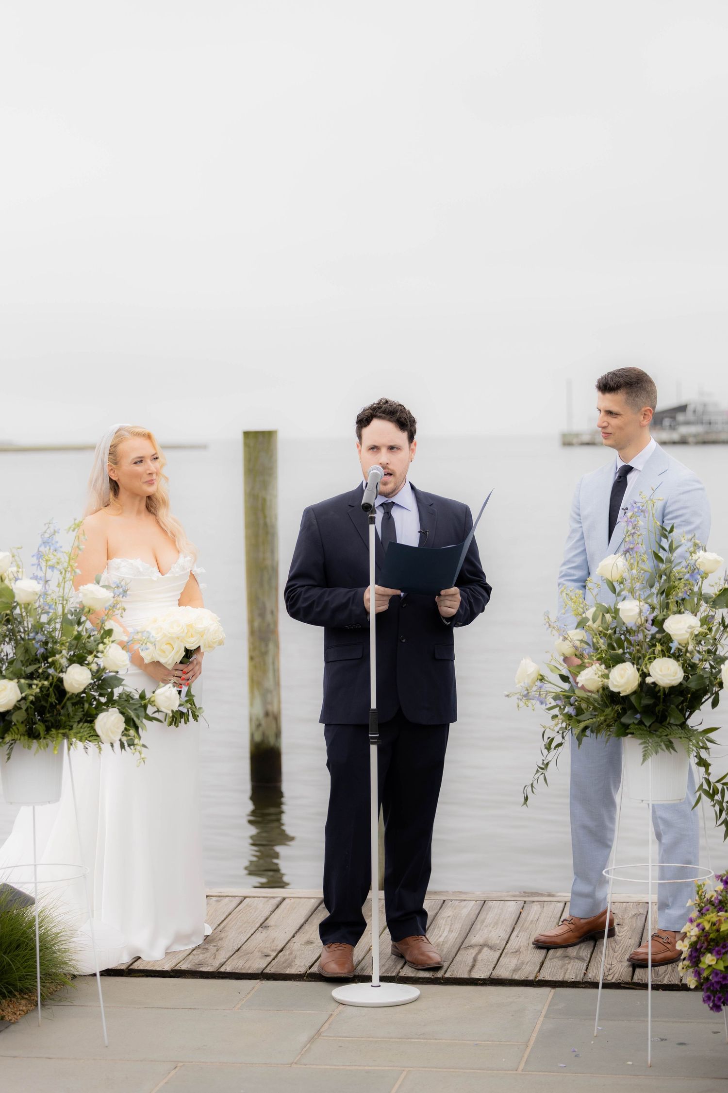 Wedding party members stand on a wooden dock by the water during a coastal celebration ceremony.