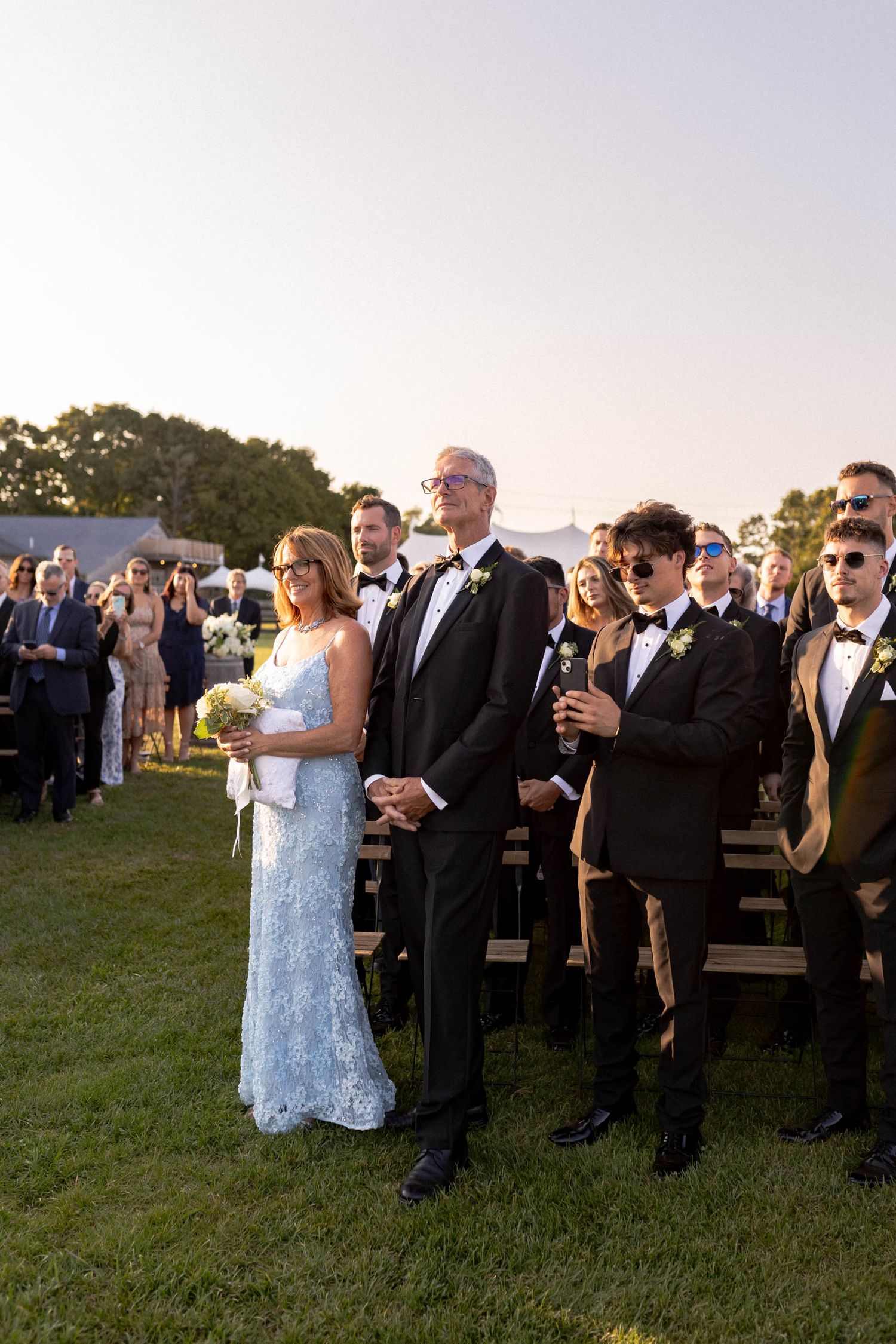 Wedding guests gather on a grassy lawn at sunset for an outdoor ceremony celebration.