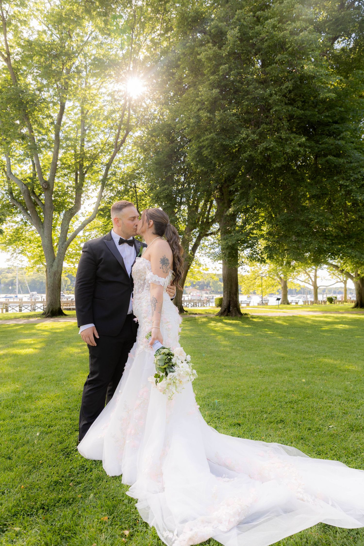 A romantic wedding couple shares a kiss in a sunlit park with lush trees and a flowing white dress train.