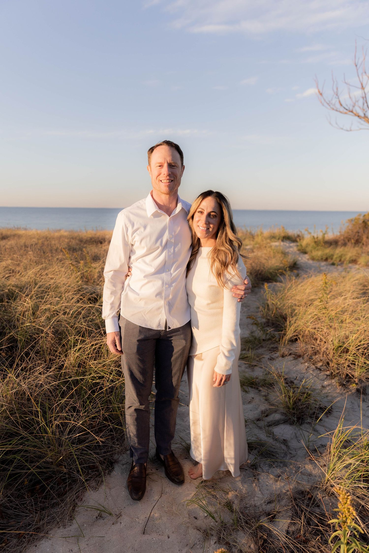 Couple poses together on sandy beach dunes at sunset with ocean in background.