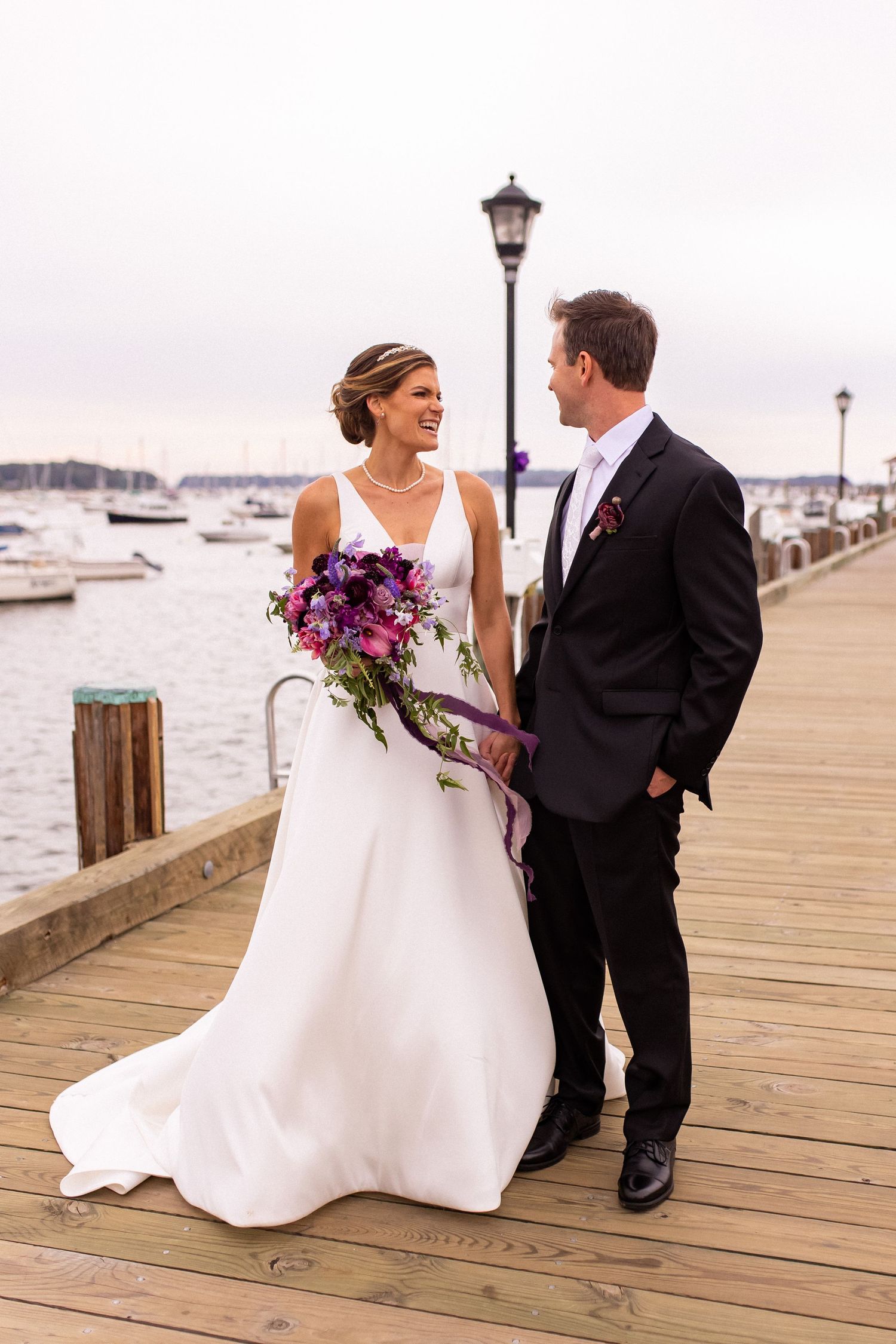 A couple in wedding attire shares a romantic moment while walking along a wooden dock with boats in the background.