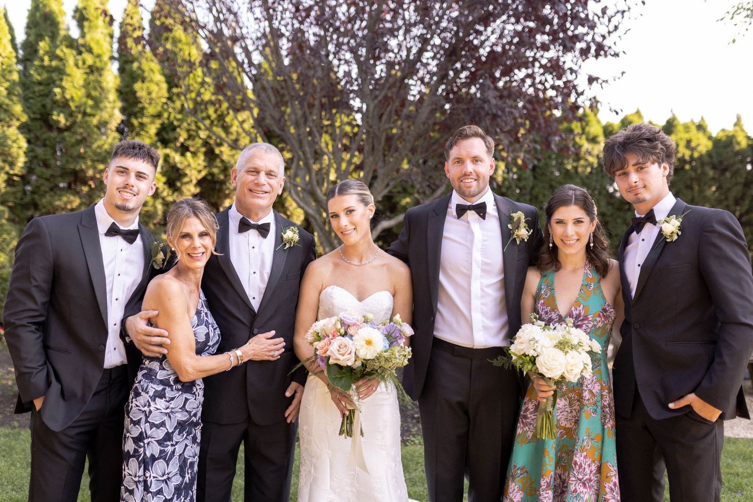 Wedding party in formal attire poses together outdoors with bouquets during sunset.