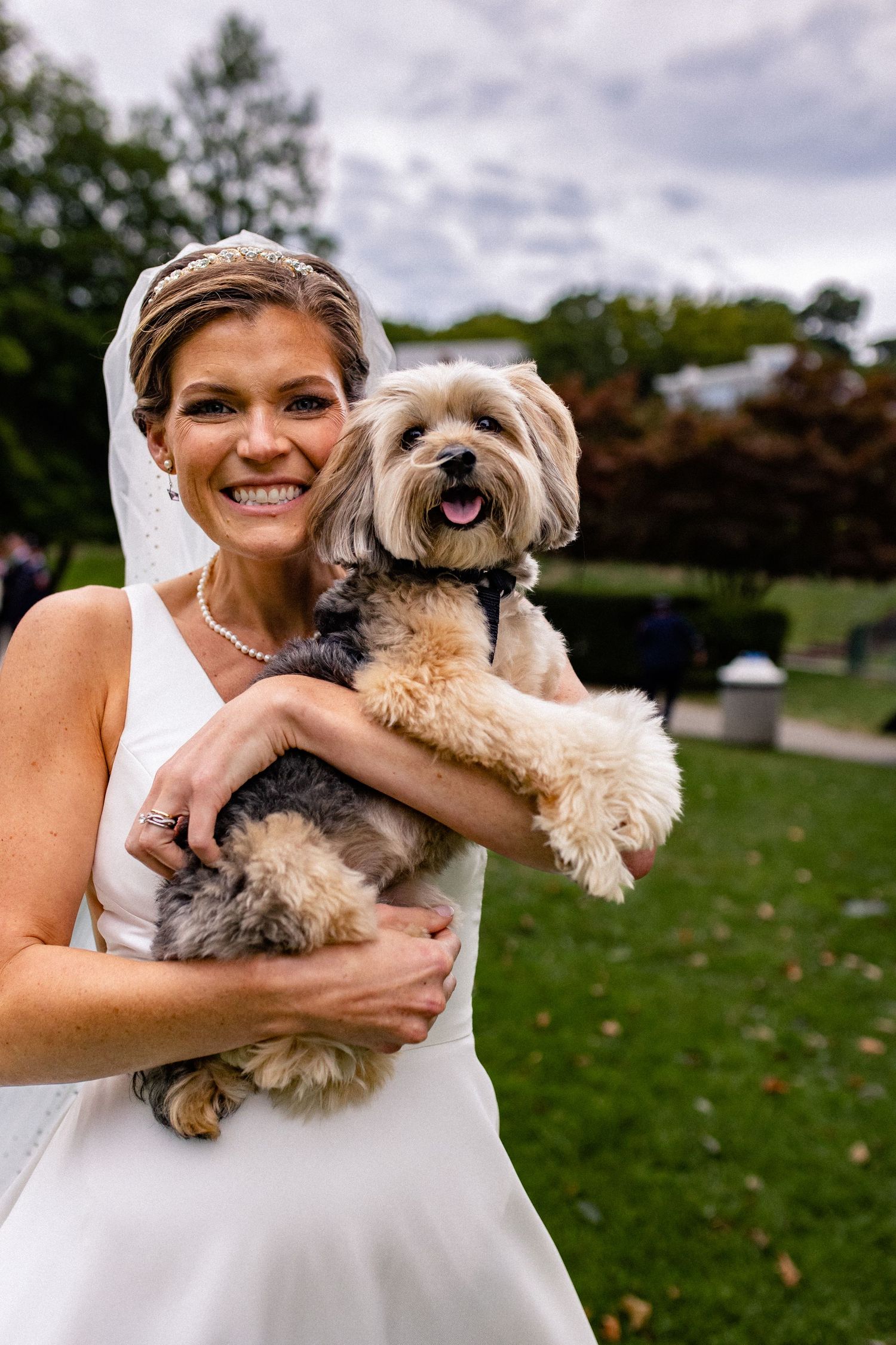 A happy bride in a white dress holds her smiling Havanese dog outdoors during a wedding photoshoot.