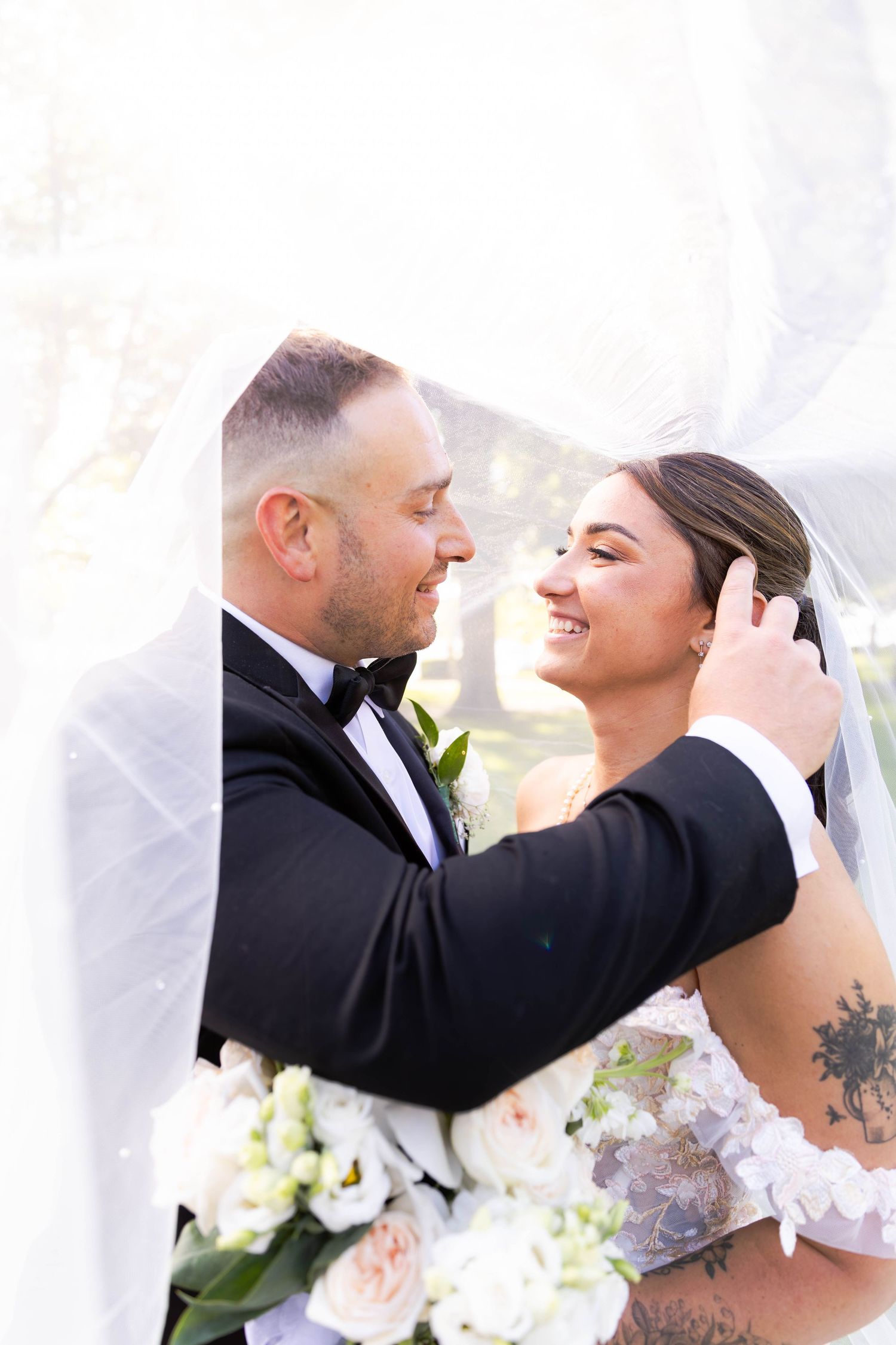 A romantic wedding moment captured between newlyweds sharing a tender embrace under a white veil with floral bouquet.