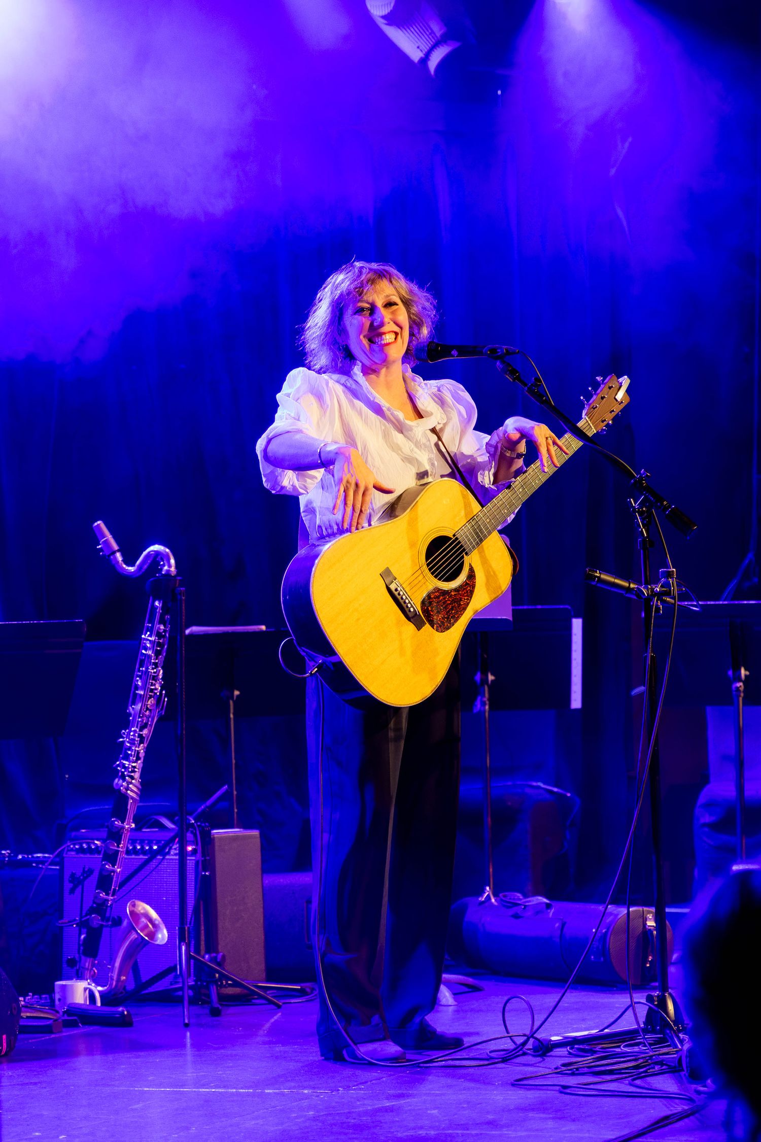 Musician performs with acoustic guitar on stage illuminated by dramatic blue and purple lighting.