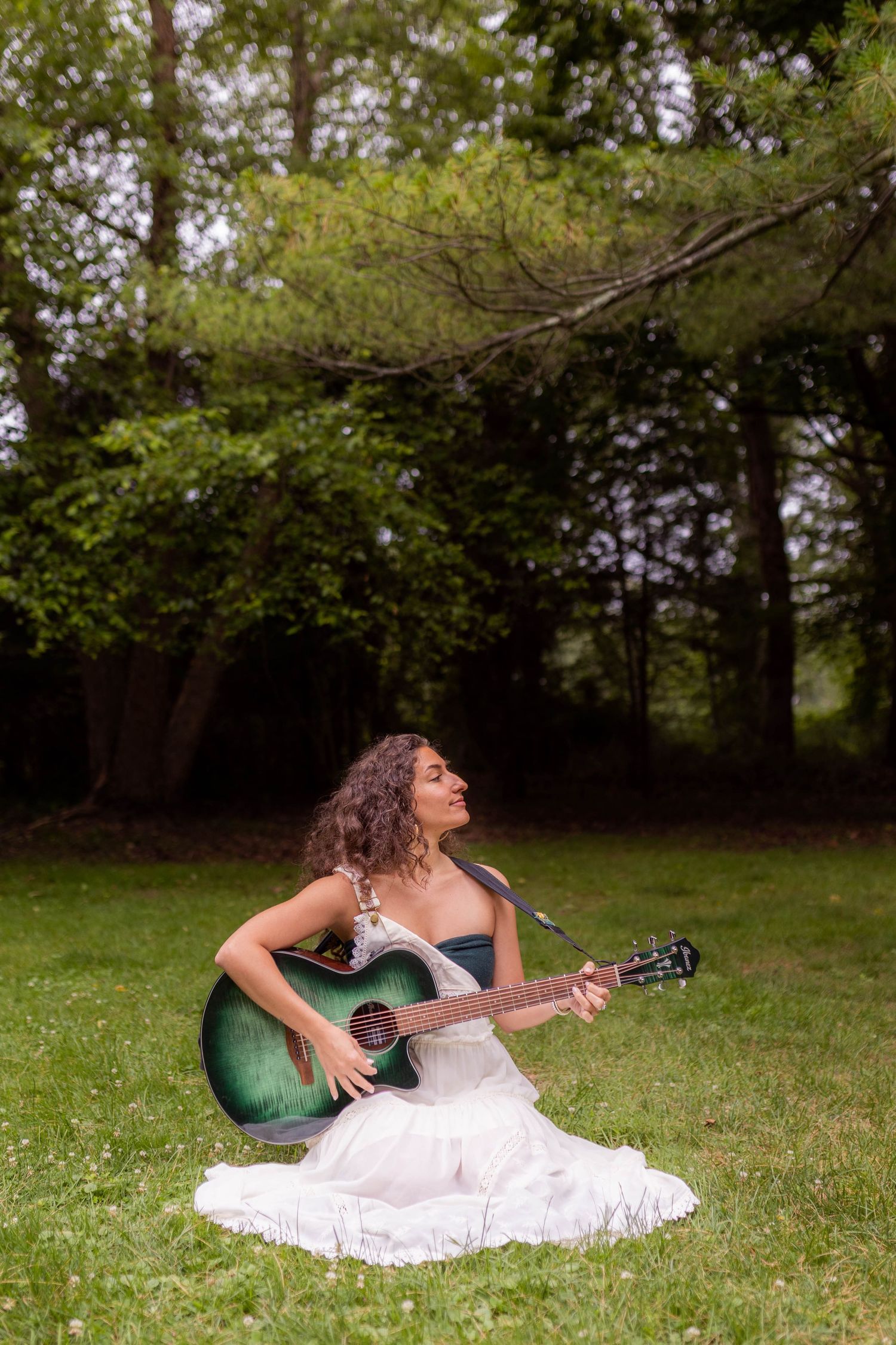 A musician in a white dress plays acoustic guitar while sitting on grass in a wooded setting.