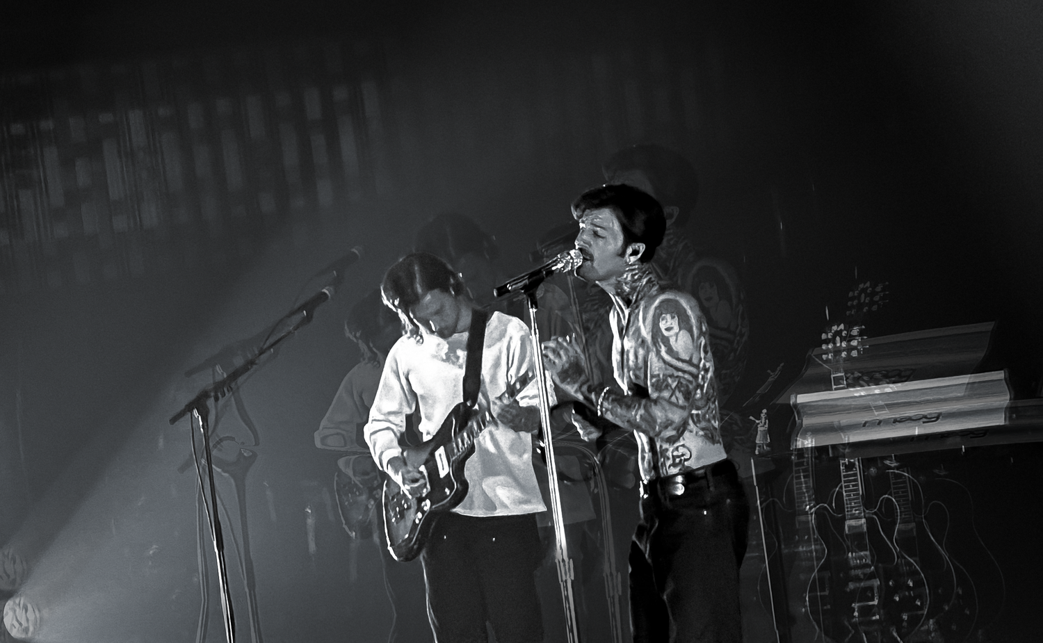 Two musicians performing on stage in black and white, one playing guitar and one singing into a microphone.