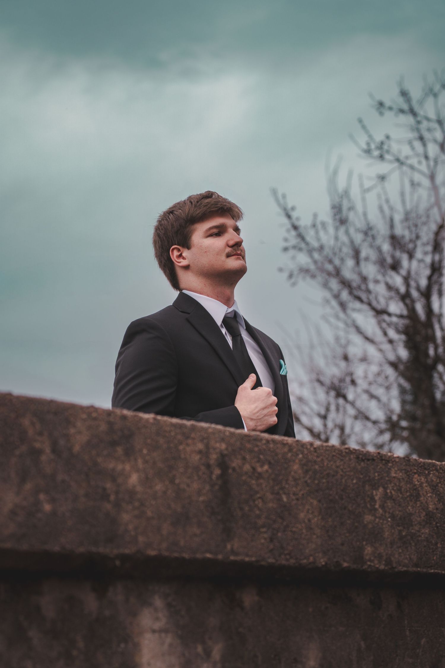 Young man in black suit and tie standing confidently outdoors against cloudy sky near stone wall.