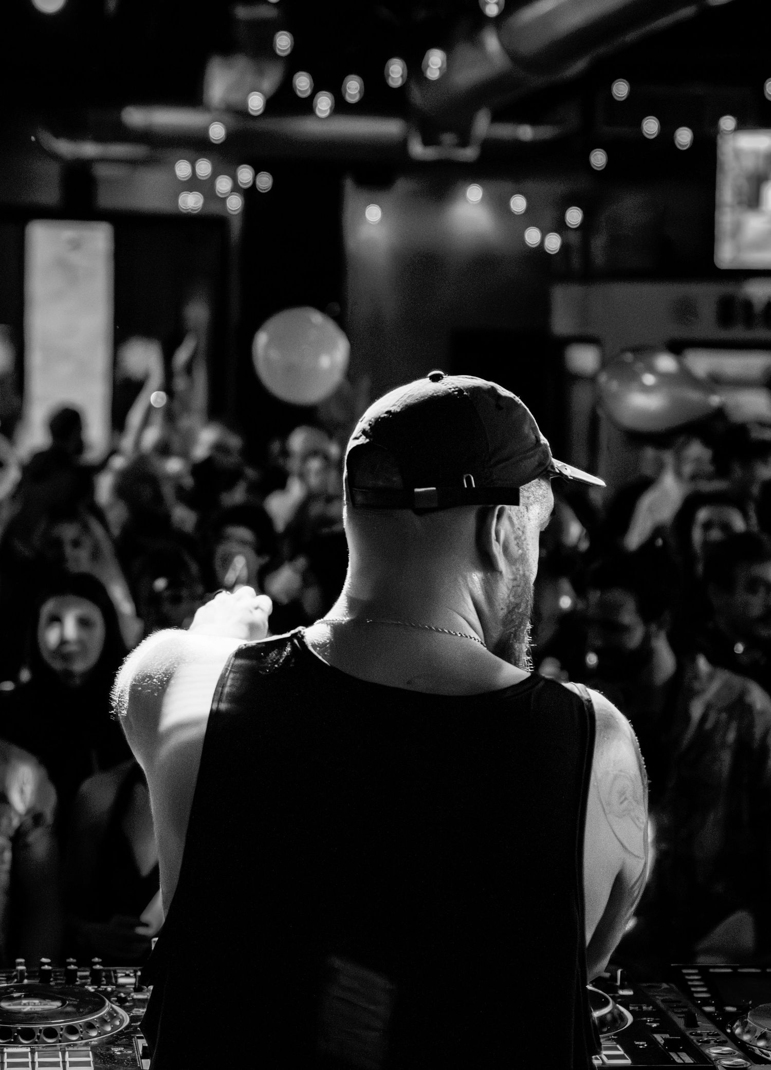 Black and white photo of a DJ performing at a crowded nightclub event, viewed from behind.
