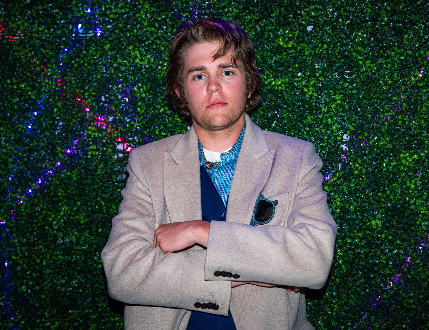 Young man in beige blazer standing with arms crossed in front of a green hedge wall with fairy lights.