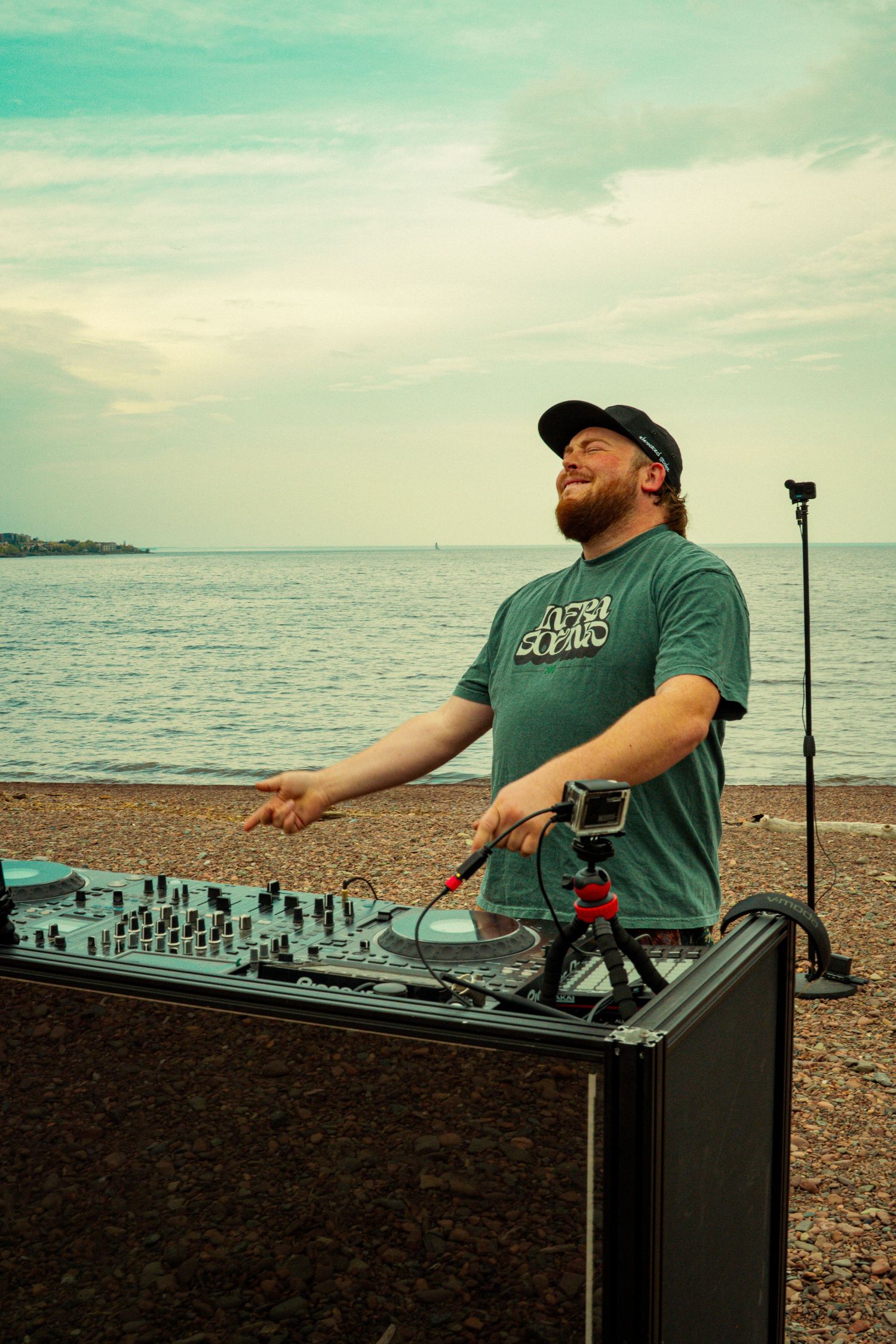 Smiling bearded DJ operating a mixer on a pebble beach with ocean backdrop on a cloudy day.