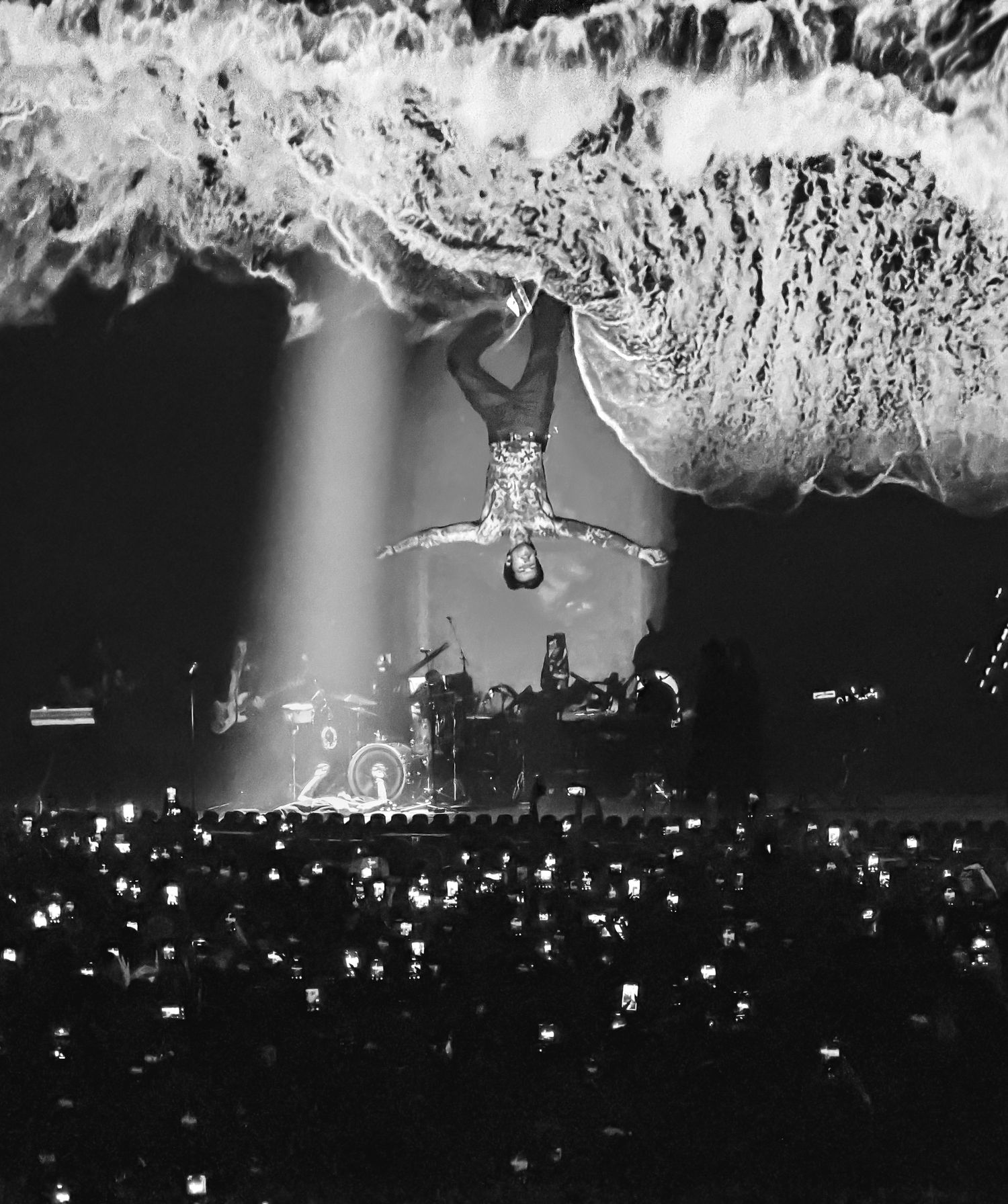 Black and white concert photo of acrobat hanging upside down above cheering crowd with dramatic light beams and sparks.