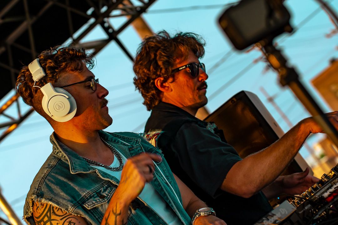 Two DJs performing at outdoor festival, one wearing white headphones, both operating mixing equipment at sunset.