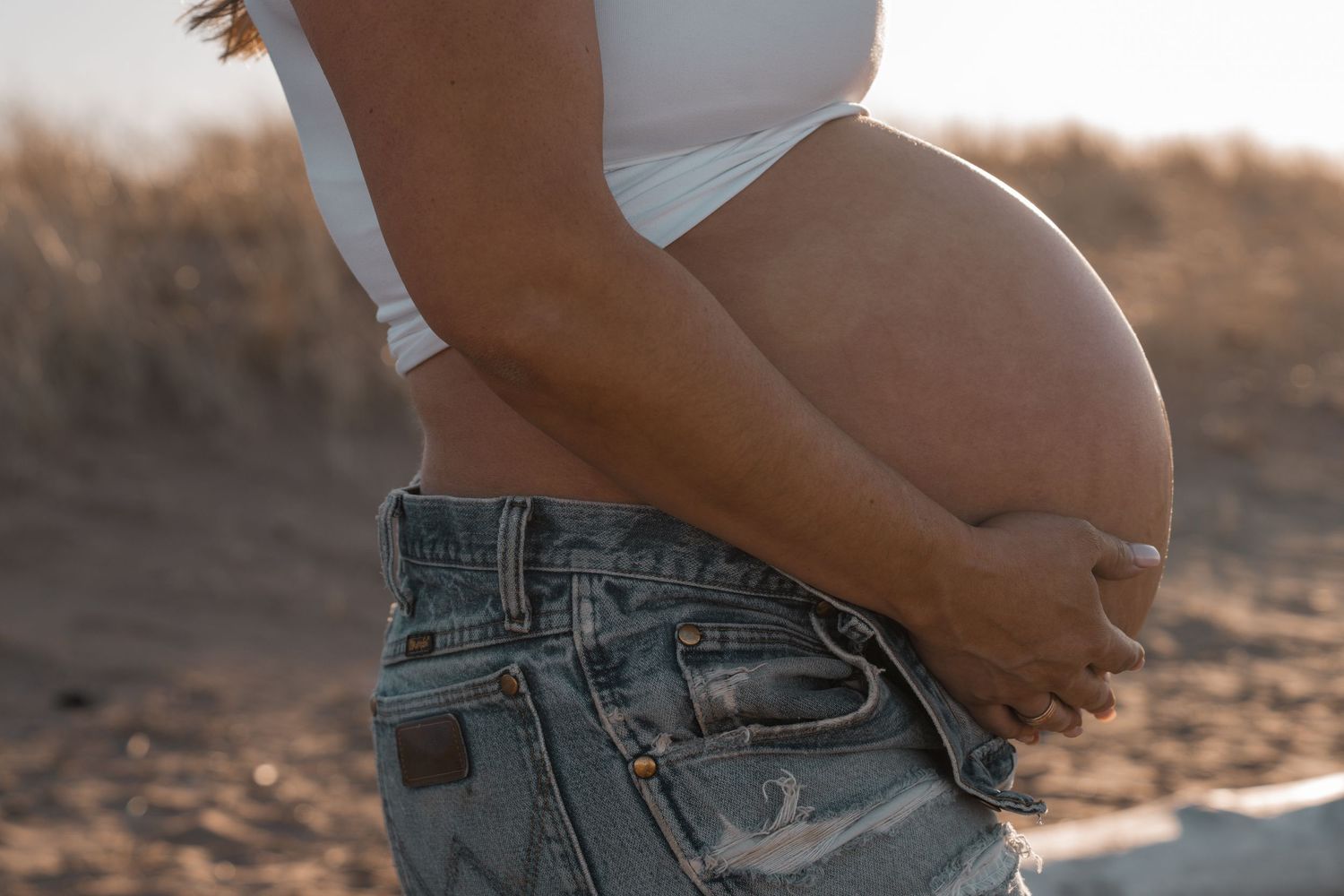 Pregnant woman in white crop top and open jeans cradling baby bump outdoors in sunny desert setting.