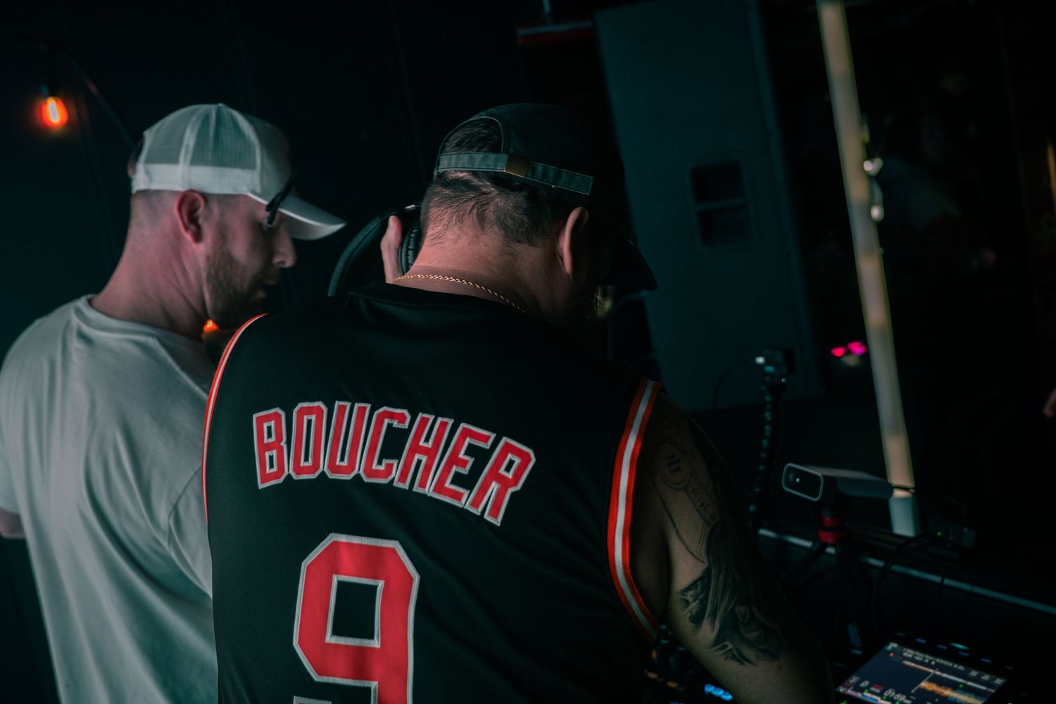 Two DJs performing at a nightclub, one wearing a Boucher number 9 jersey, working DJ equipment in dark venue.